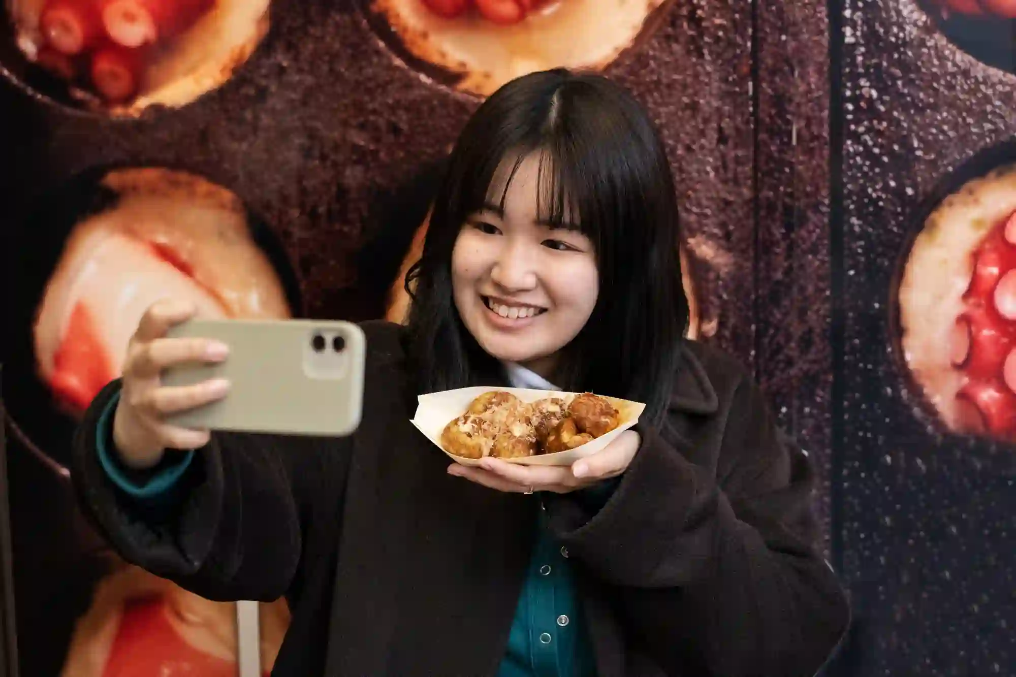 A woman takes a selfie while holding a plate of takoyaki in front of a large octopus snack mural.