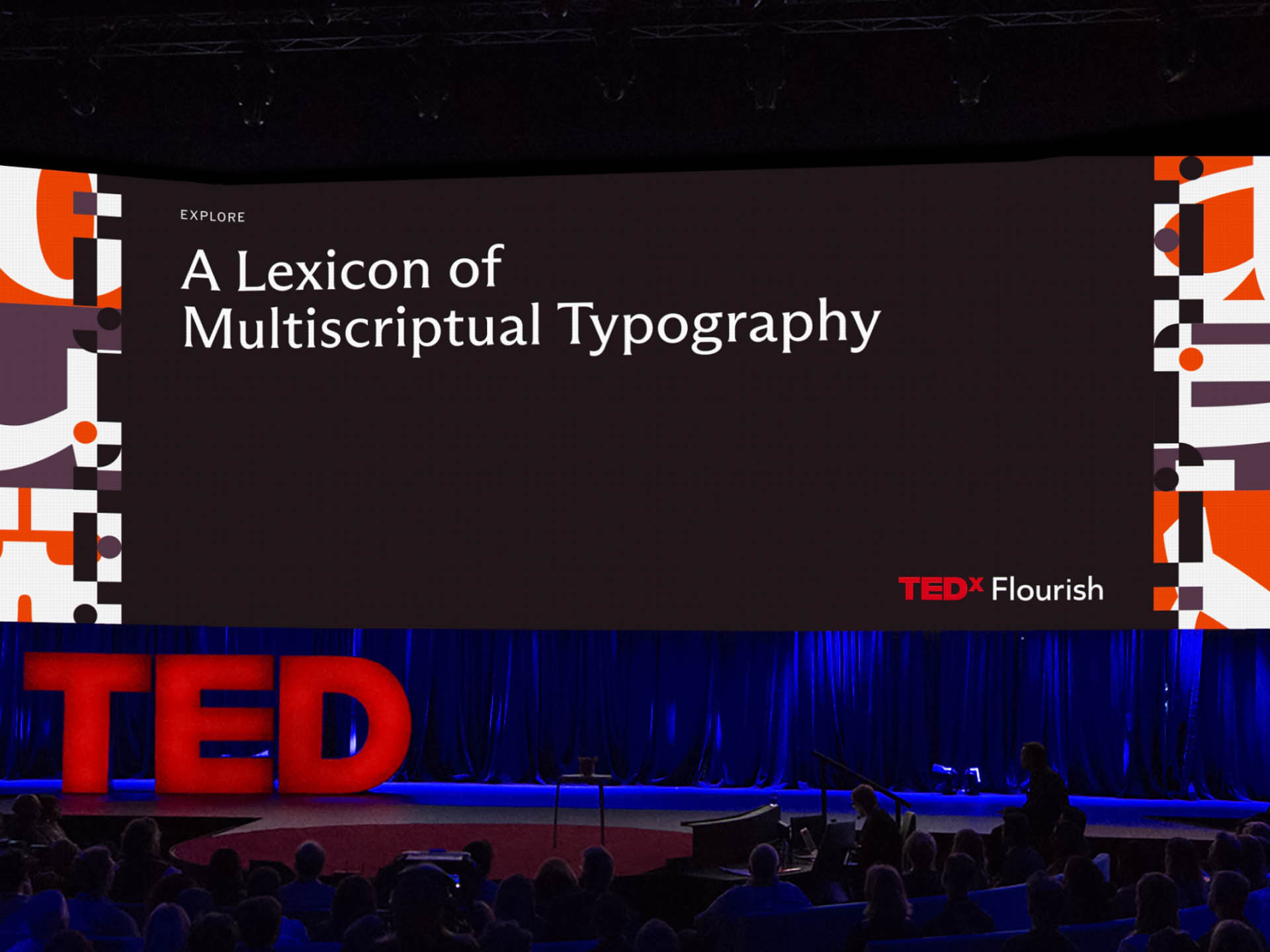 A presentation slide titled “A Lexicon of Multiscriptual Typography” is displayed on a dark screen. The TEDx Flourish logo is visible in the bottom right corner. The slide features geometric shapes and text in white and orange. A stage with blue lighting and audience members is visible below the screen.