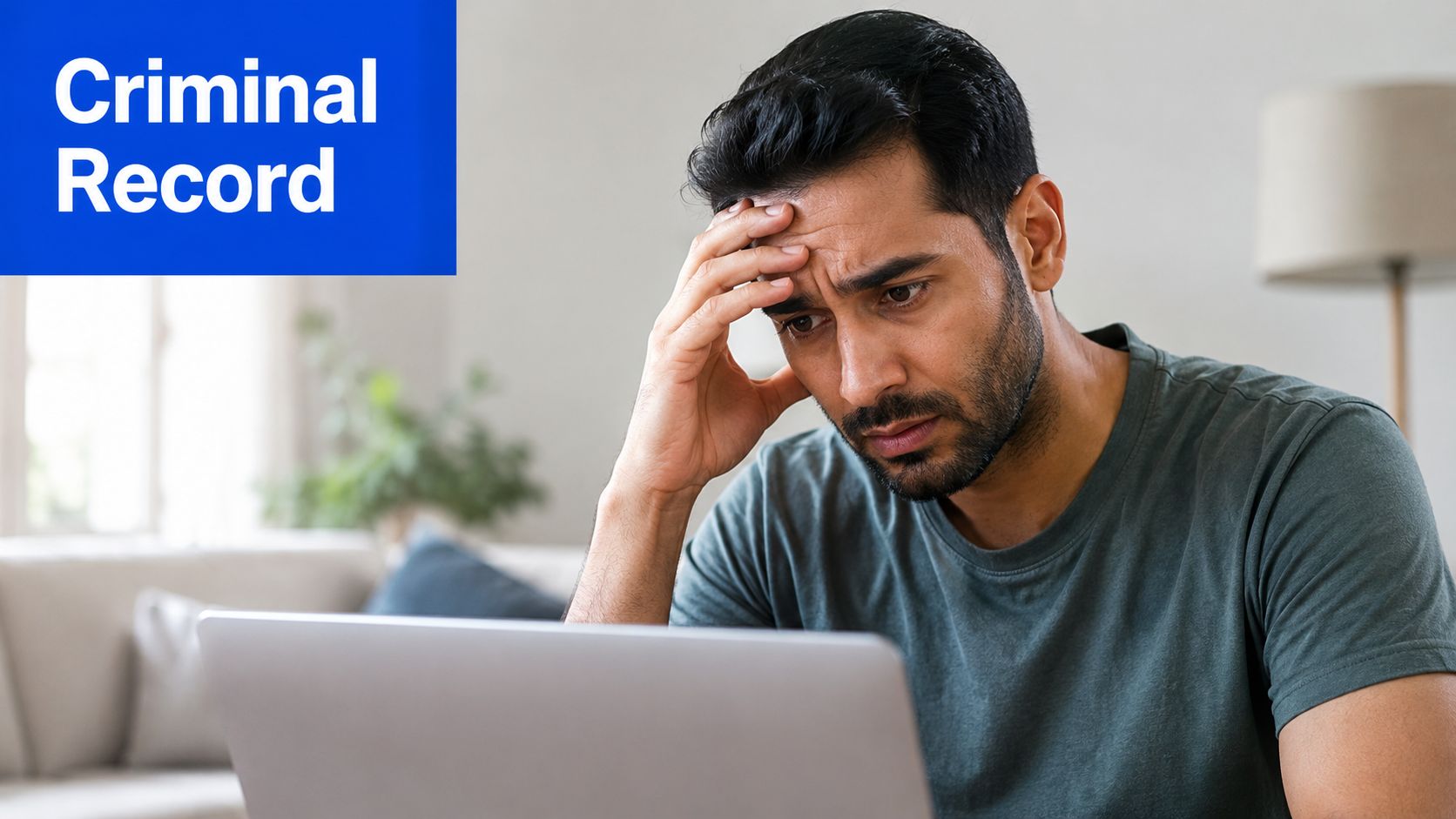 A concerned man sits at a laptop looking stressed while researching information about a criminal record.