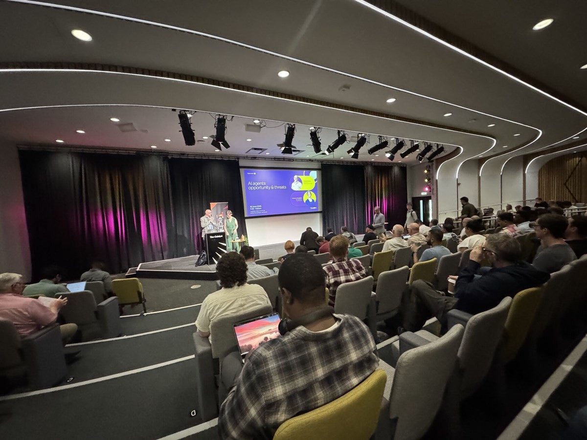 Audience seated in a modern auditorium watching two presenters on stage in front of a large screen displaying a slide titled ‘AI agents: opportunity & threats.’ The stage has a podium and colorful lighting.