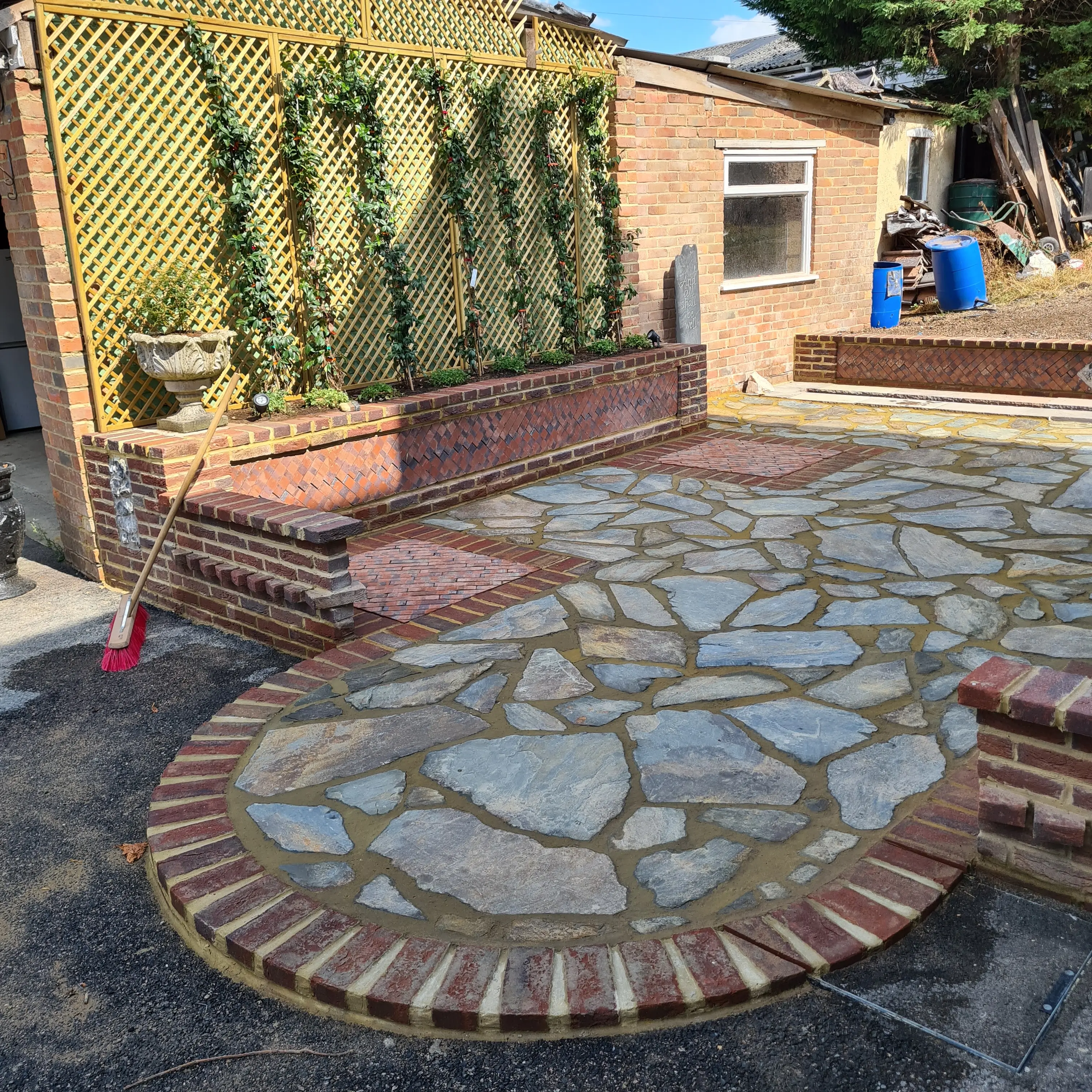 A newly laid stone patio with a circular design, surrounded by greenery and a building in the background.