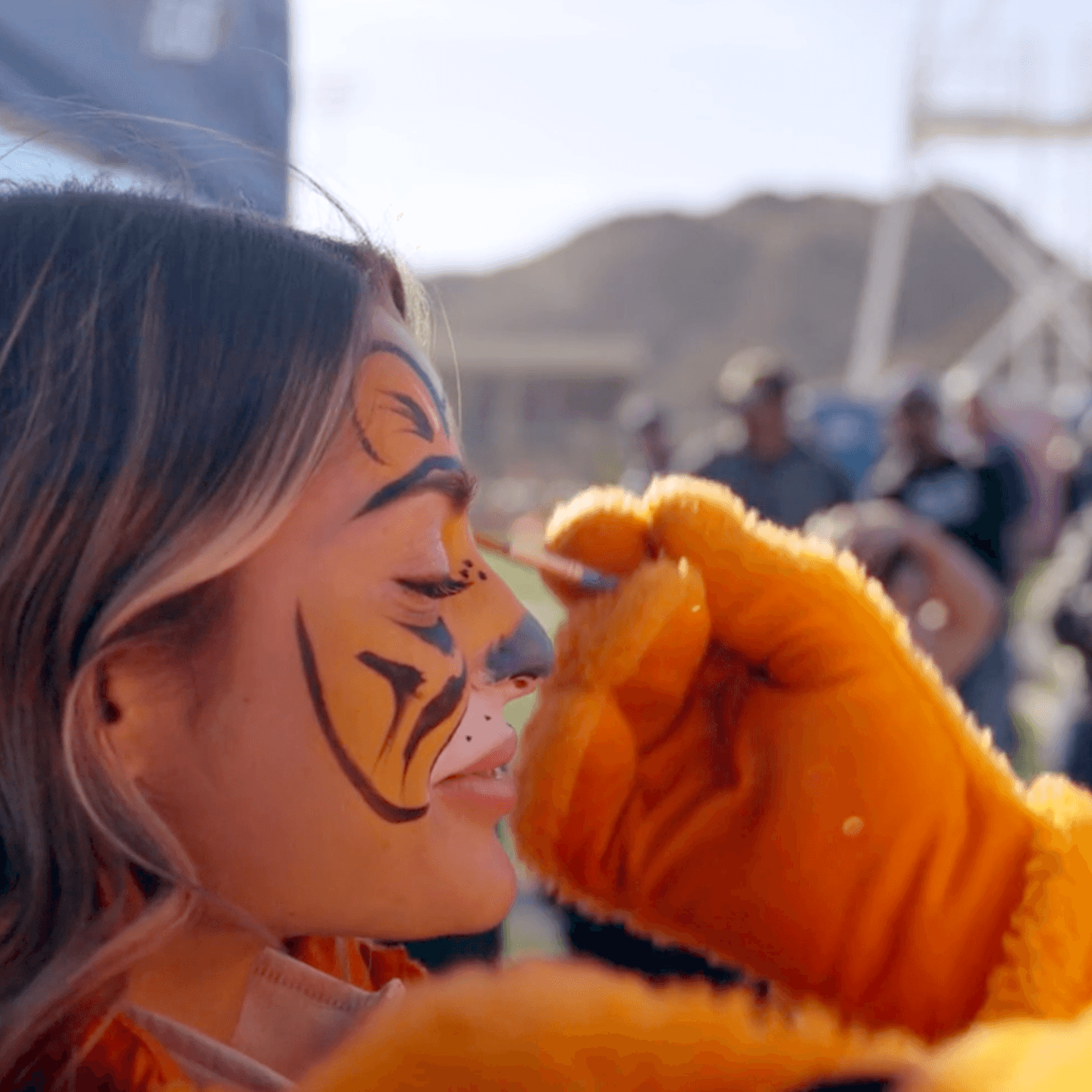 A person wearing orange gloves paints tiger stripes on a smiling womans face at an outdoor event, with blurred people and hills in the background.