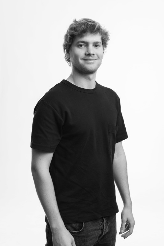 Black-and-white studio portrait of a smiling man with short curly hair and light stubble, wearing a black short-sleeve pocket t-shirt and jeans, standing in a three-quarter pose against a plain light background.