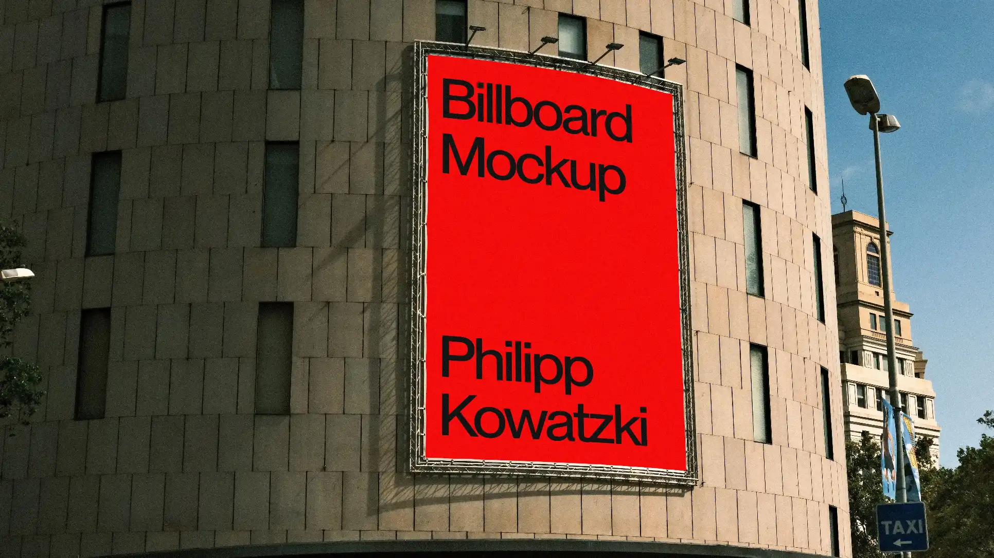 A large rectangular billboard mockup mounted on a modern curved building facade with a tiled exterior. The setting includes urban surroundings, trees, and a clear blue sky with streetlights.