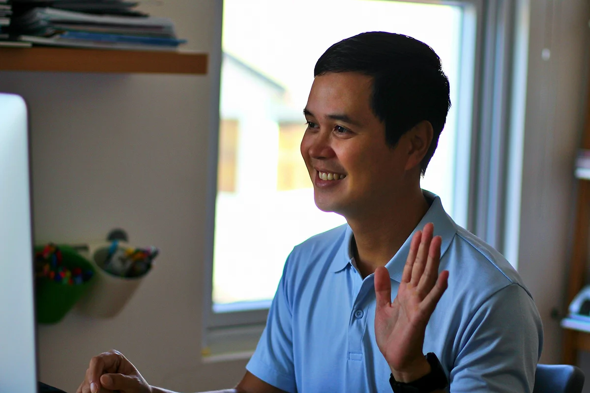 A smiling man waving during a video call at his laptop.