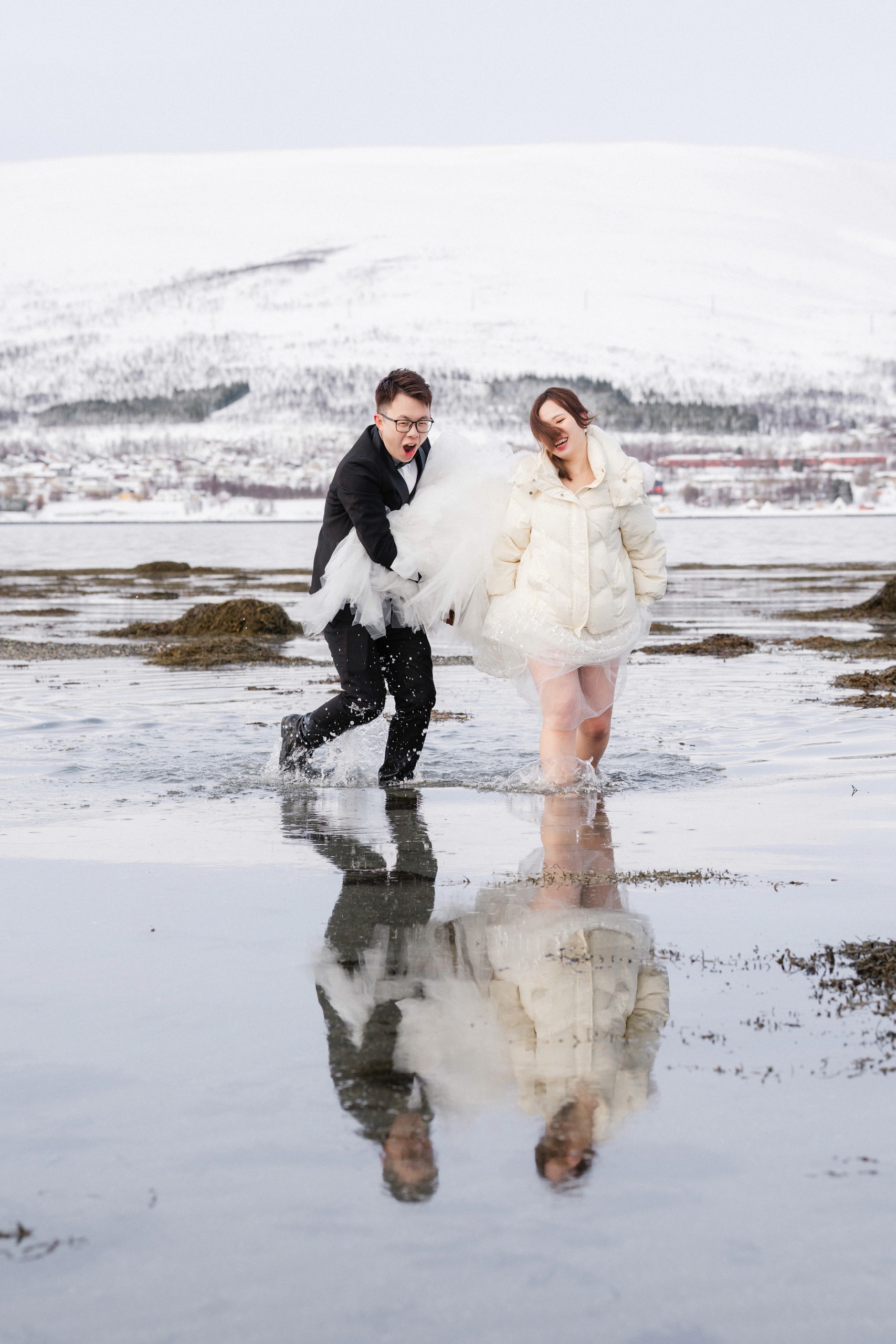 The couple moves playfully through shallow shoreline water, their figures doubled in a clear reflection beneath a snowy hillside.