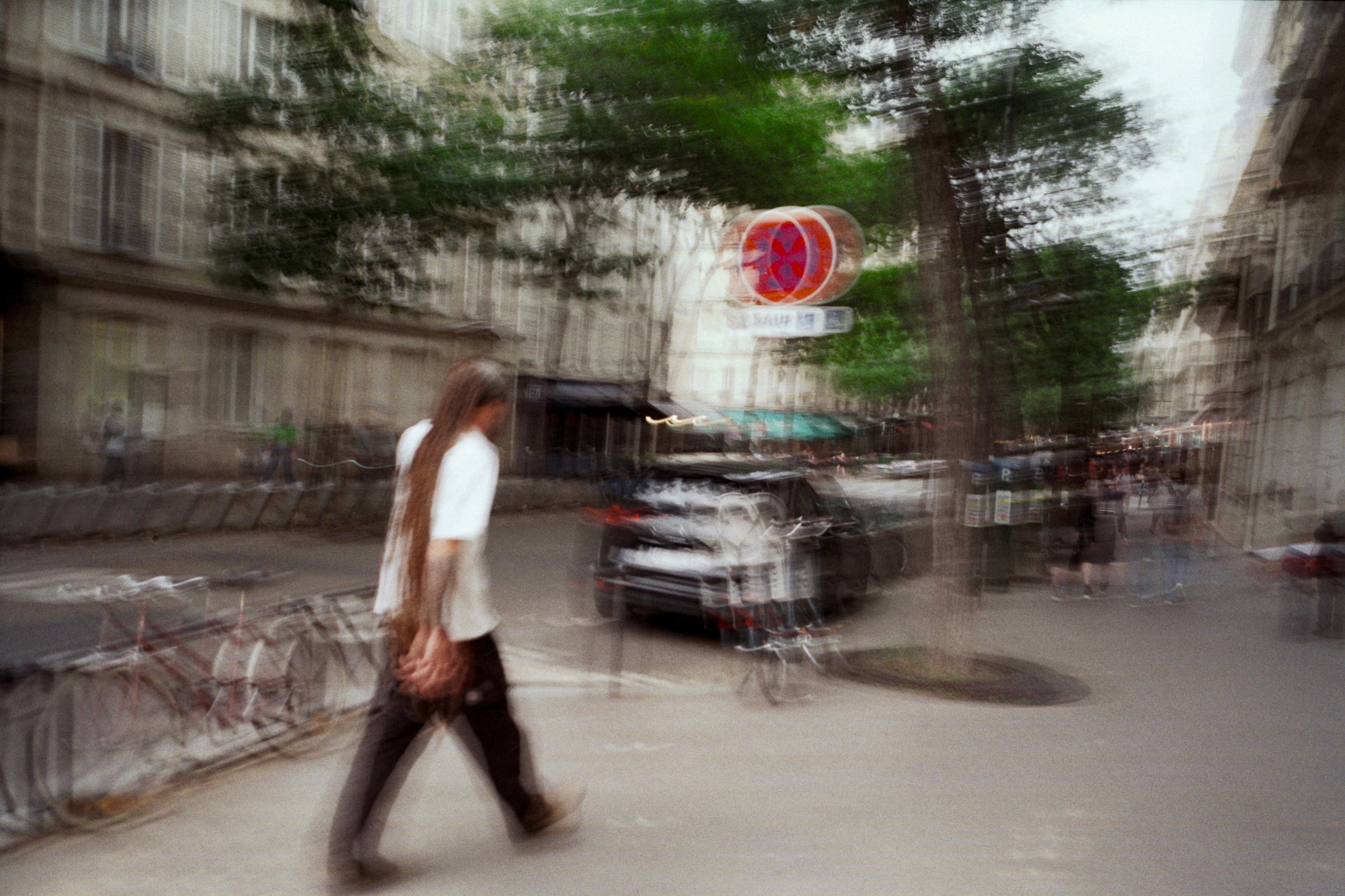 A person with long hair walks along a bustling urban street, lined with bicycles and blurred movement, under green-leaved trees and beside parked cars, capturing the vibrant, dynamic essence of city life.