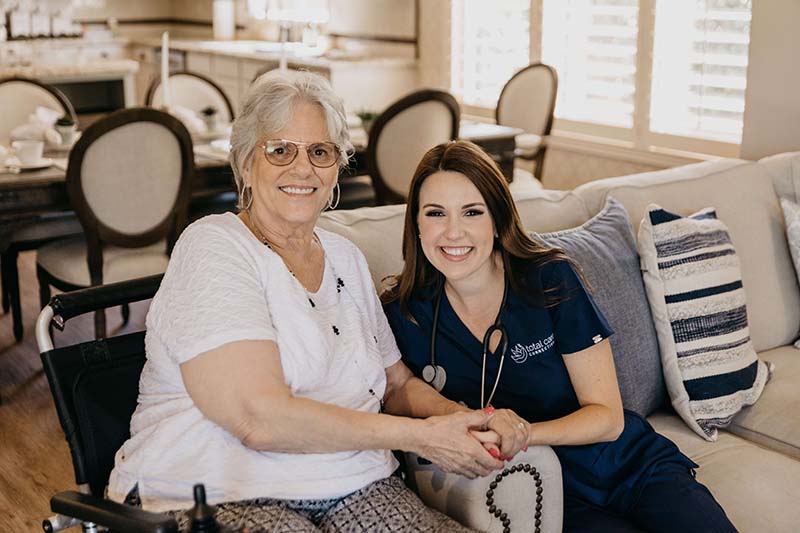 Two women sitting on a couch, one in a white shirt and the other in blue scrubs with a stethoscope around her neck, holding hands and smiling.