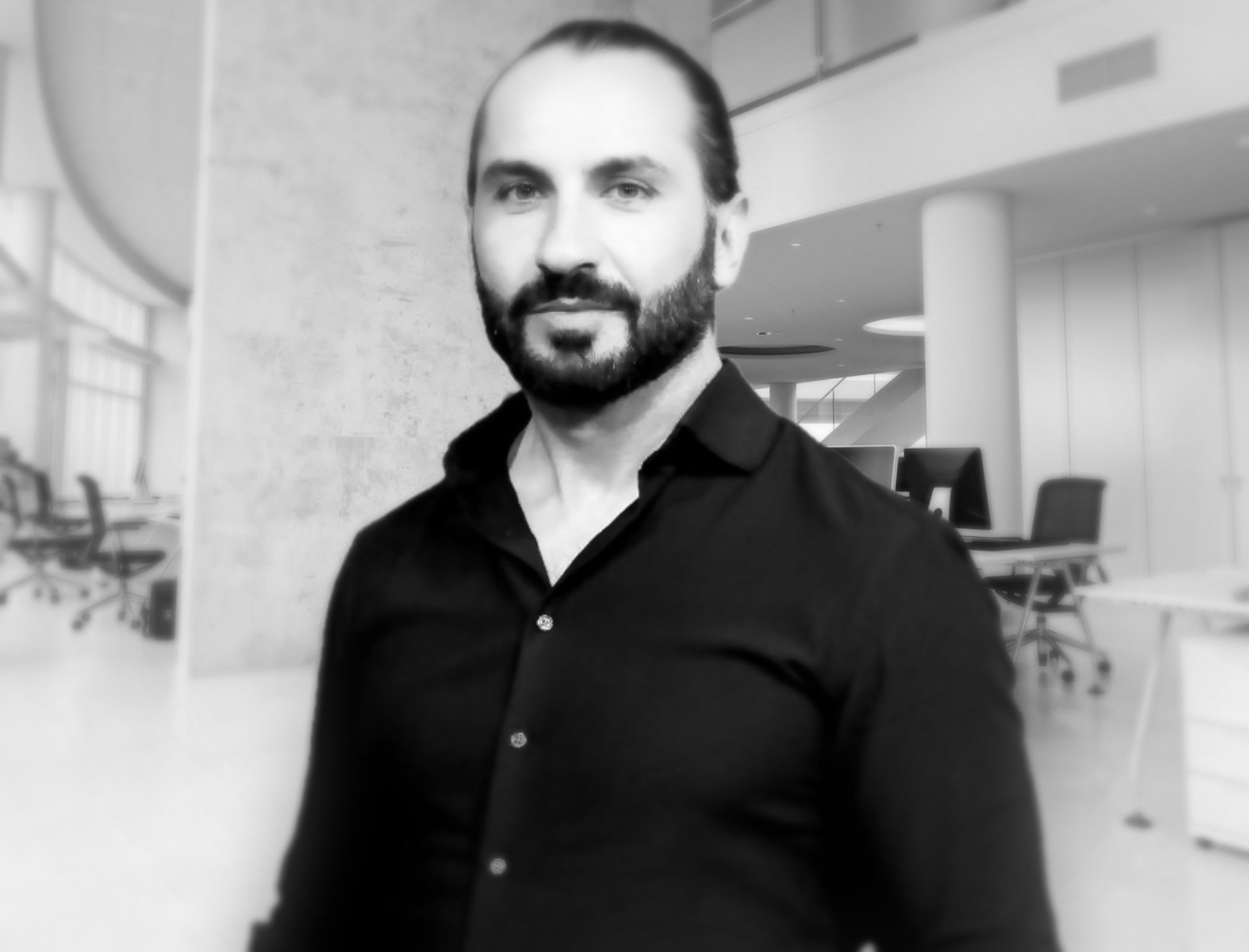 Black and white photo of a caucazian man with beard in a black shirt in a modern office interior