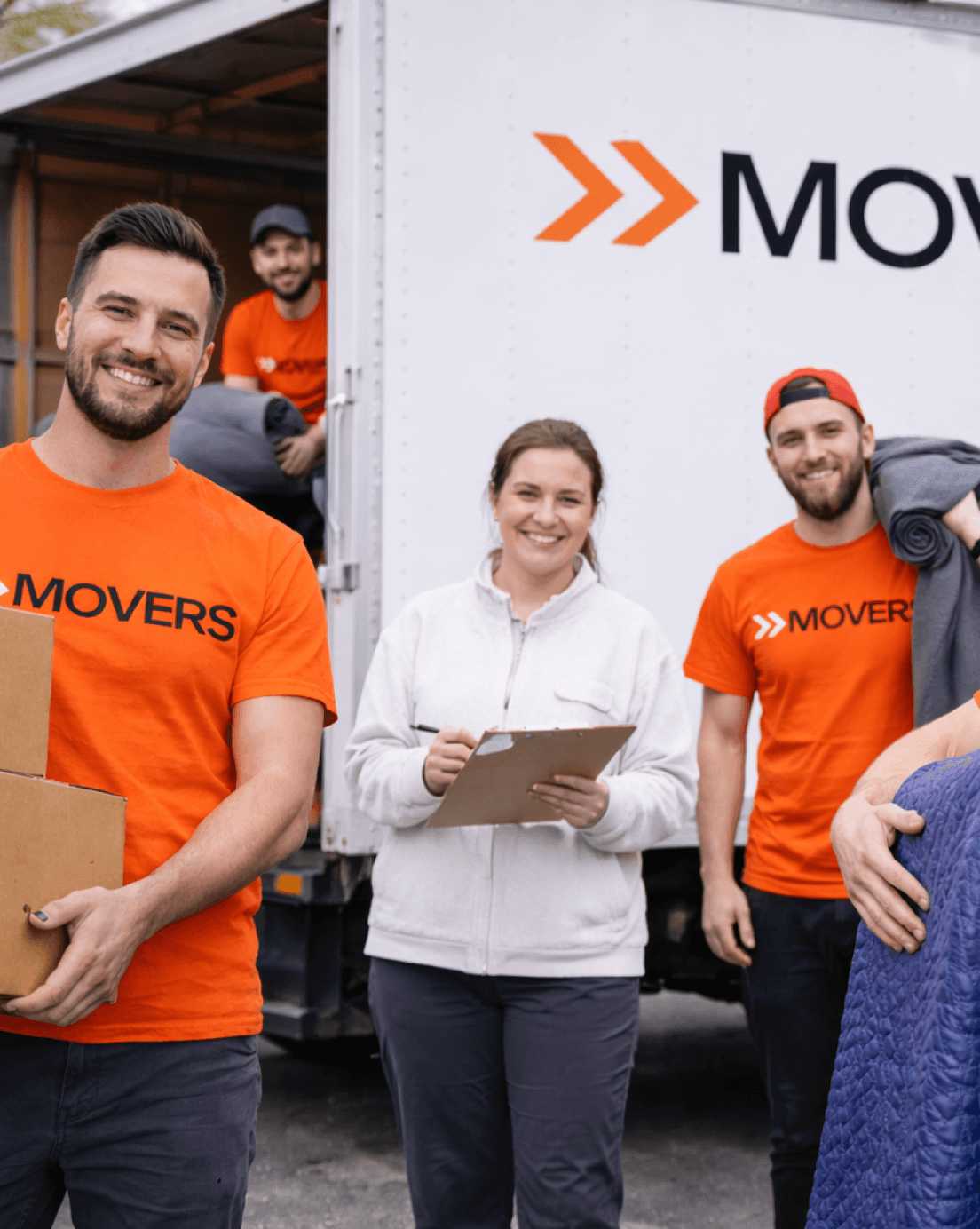 Smiling professional movers in orange t-shirts and a woman with a clipboard standing by a moving truck.