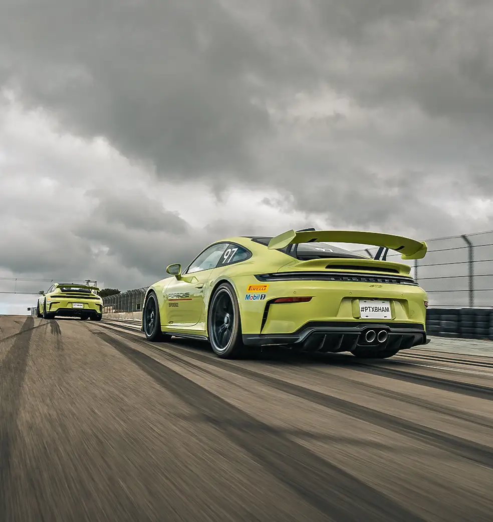 Low-angle view of yellow Porsche track cars driving on a racetrack under dark, stormy clouds.