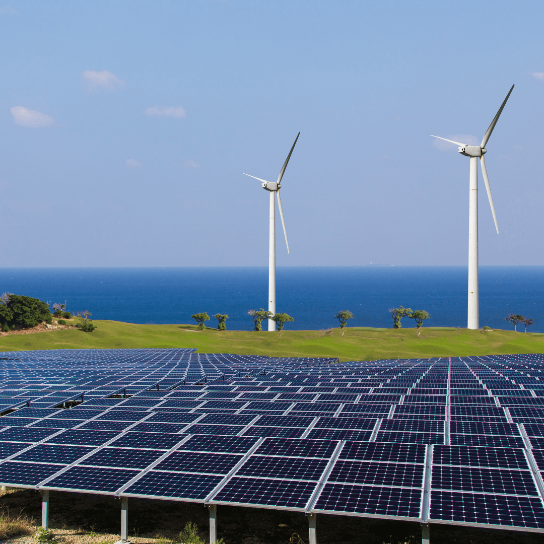 solar panels in foreground and two wind turbines on a green field, with blue sea and sky in the distance