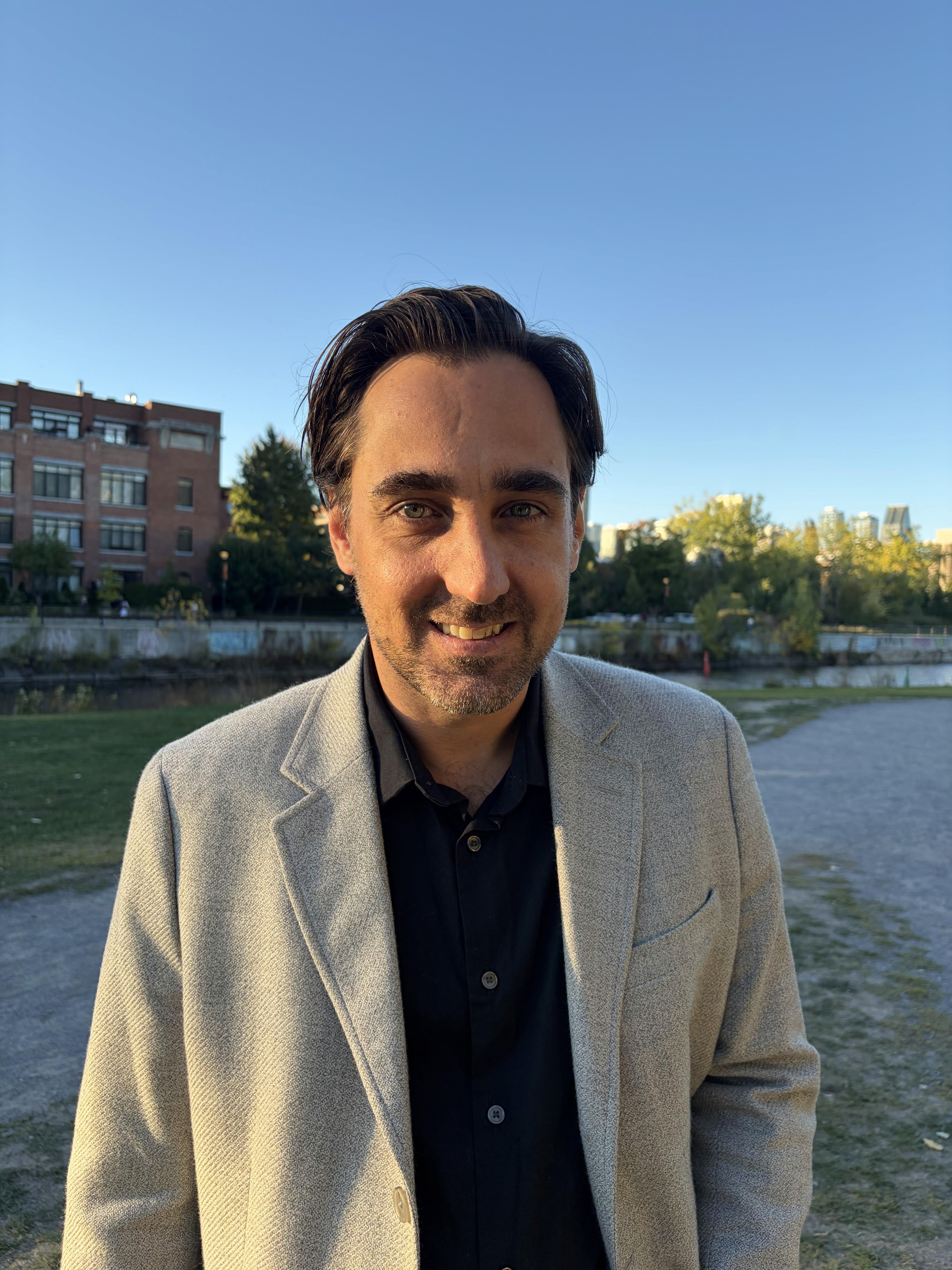 A smiling man with short hair stands with arms crossed, wearing a dark shirt against a neutral background.