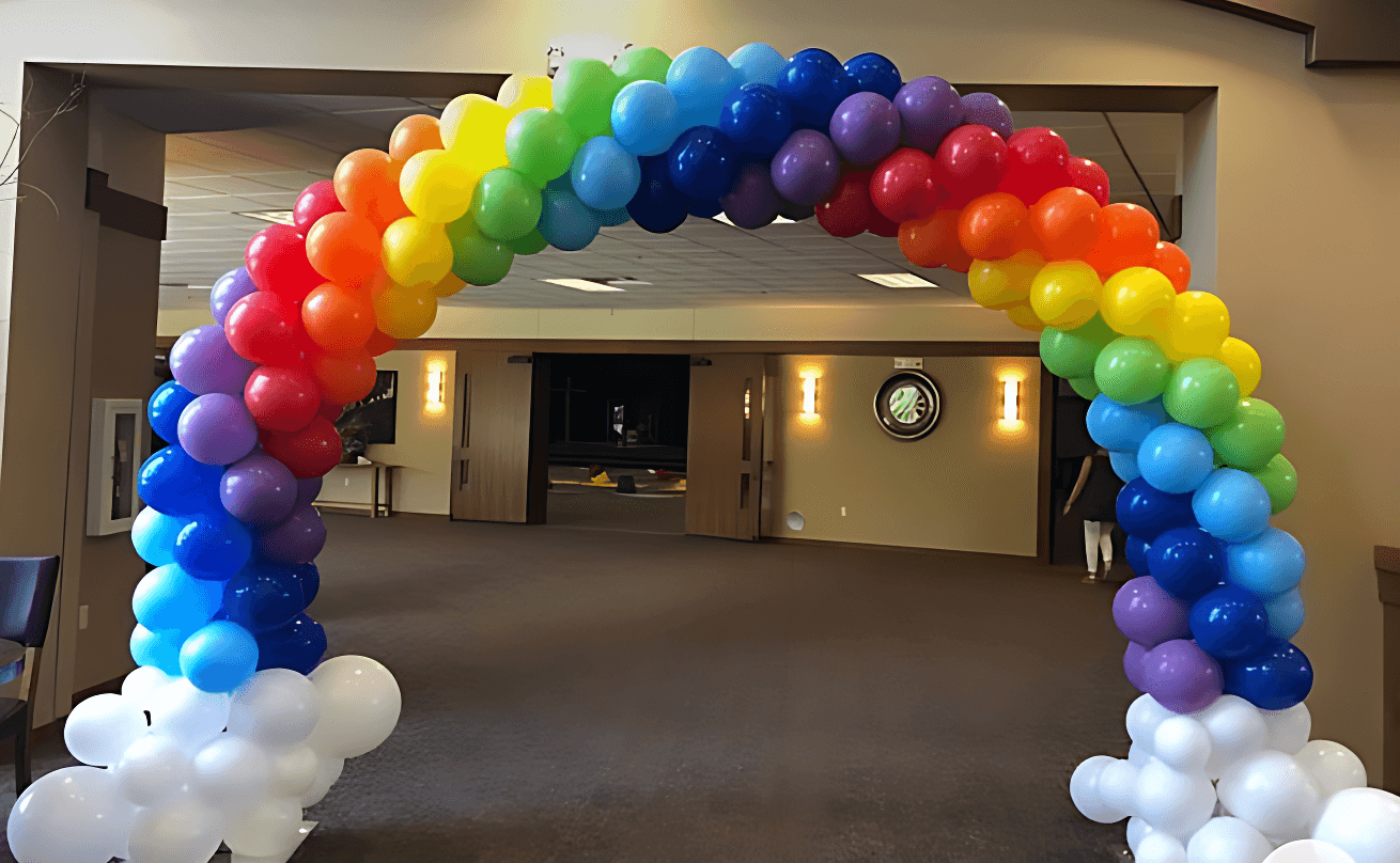A colorful balloon arch in a rainbow pattern decorates a spacious room with soft lighting and modern decor, perfect for a celebratory event.