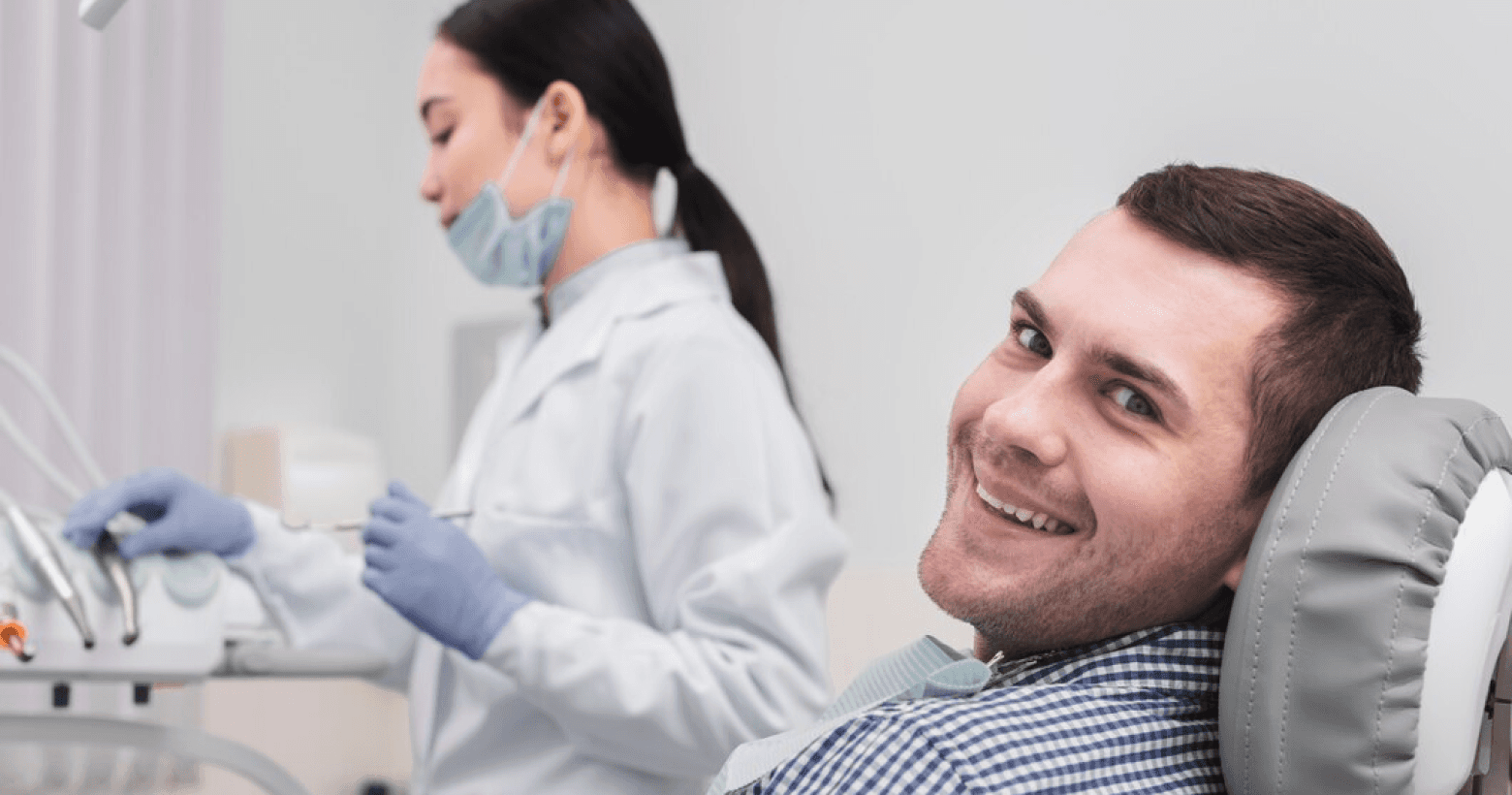 A male patient smiling while sitting in a dental chair, with a dental professional holding dental tools nearby.