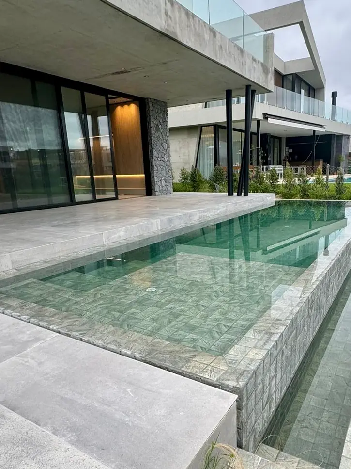 Moderna piscina elevada de diseño arquitectónico revestida en Piedra Bali, contrastando con pisos de hormigón y una casa de estilo minimalista.