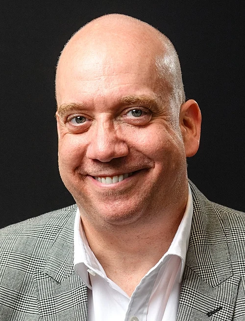 A person with a bald head and a warm smile is wearing a grey plaid blazer over a white shirt, set against a plain black background.