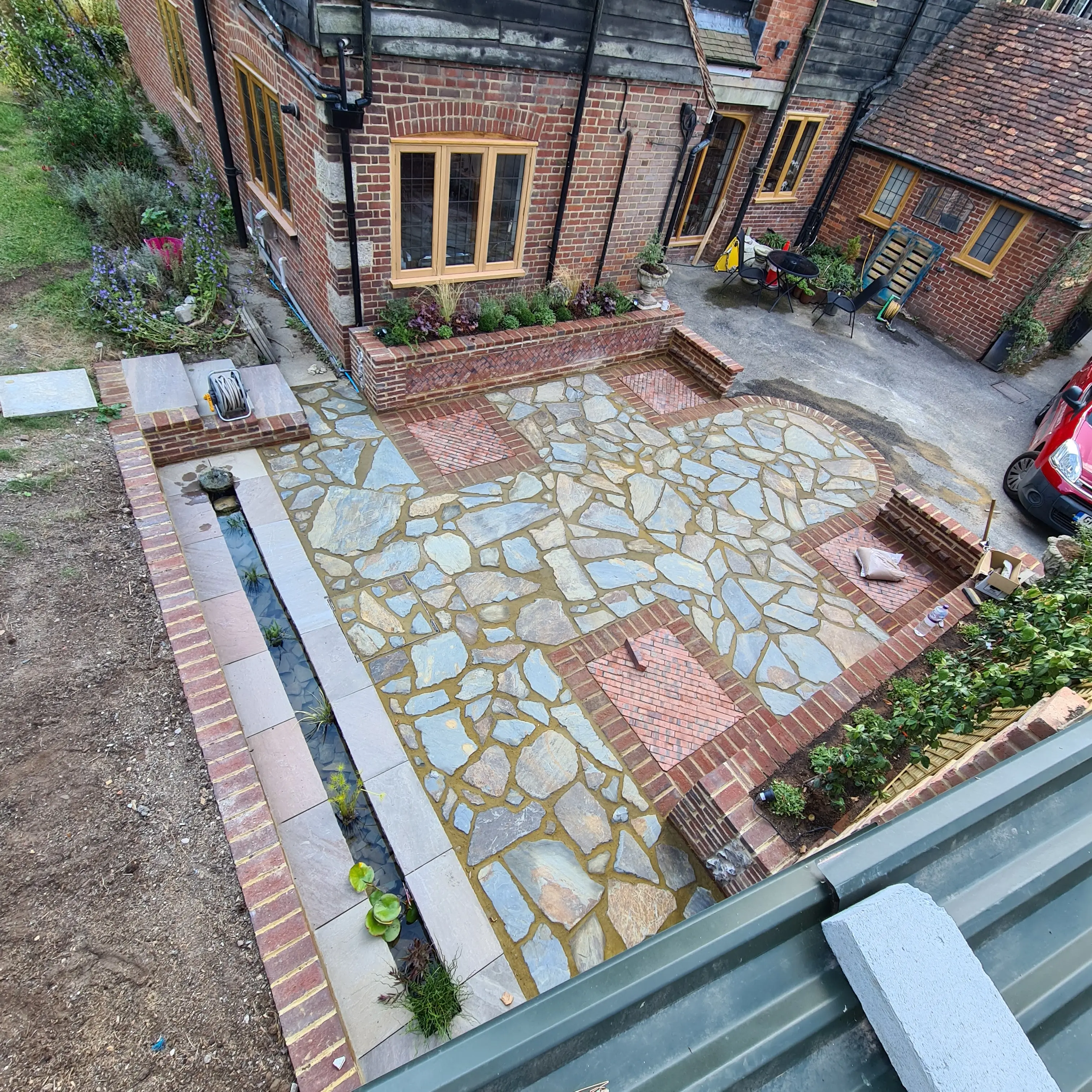 Aerial view of a landscaped patio featuring stone and brick patterns, with flower beds and a parked car nearby.