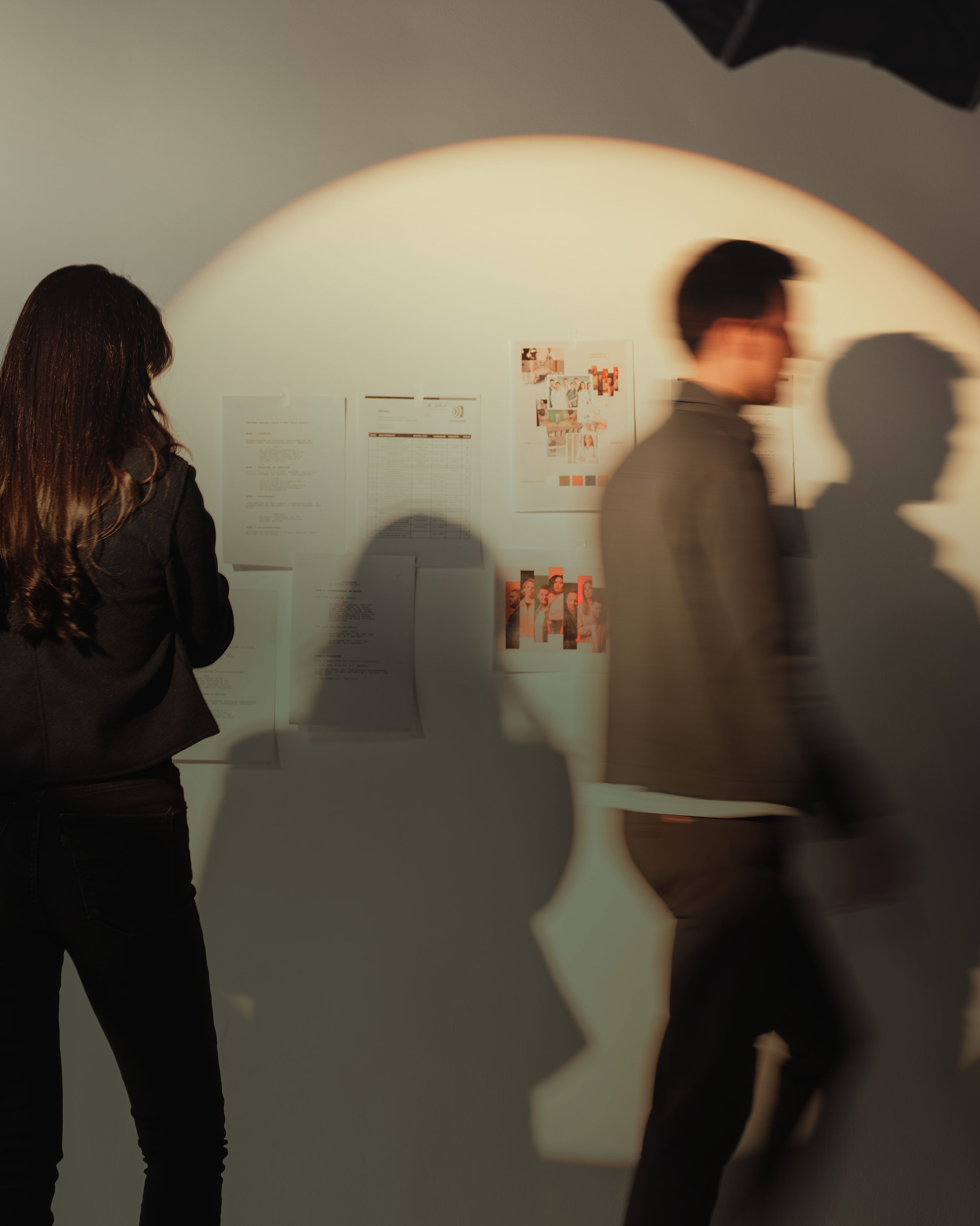 Two figures blurred in motion near a wall of papers and photographs.