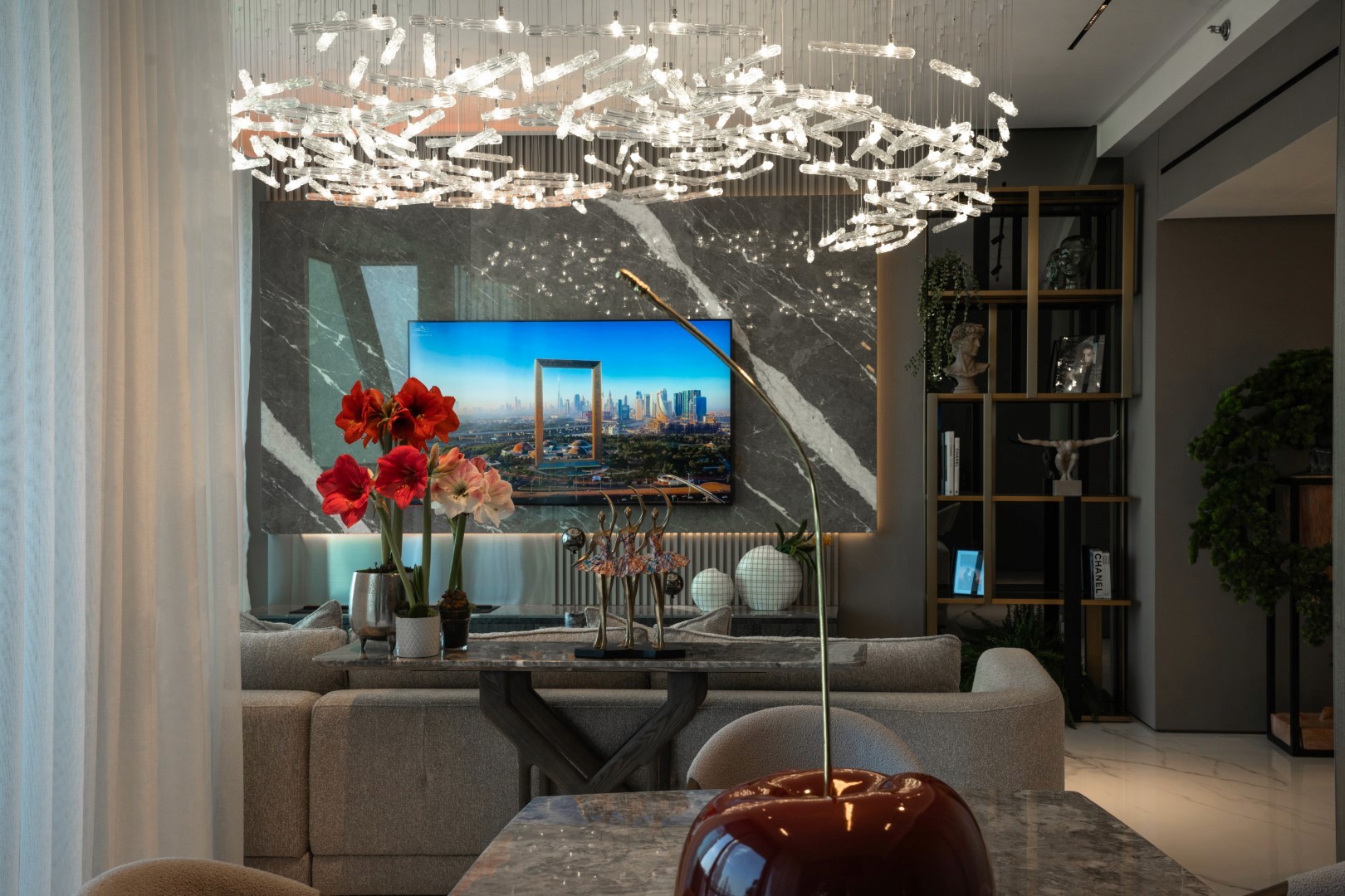 A modern living room in Burj vista, Dubai features a stylish gray couch, a sleek marble wall with a flat-screen TV displaying a cityscape, vibrant red flowers on a glass coffee table, and a unique, artistic chandelier overhead.