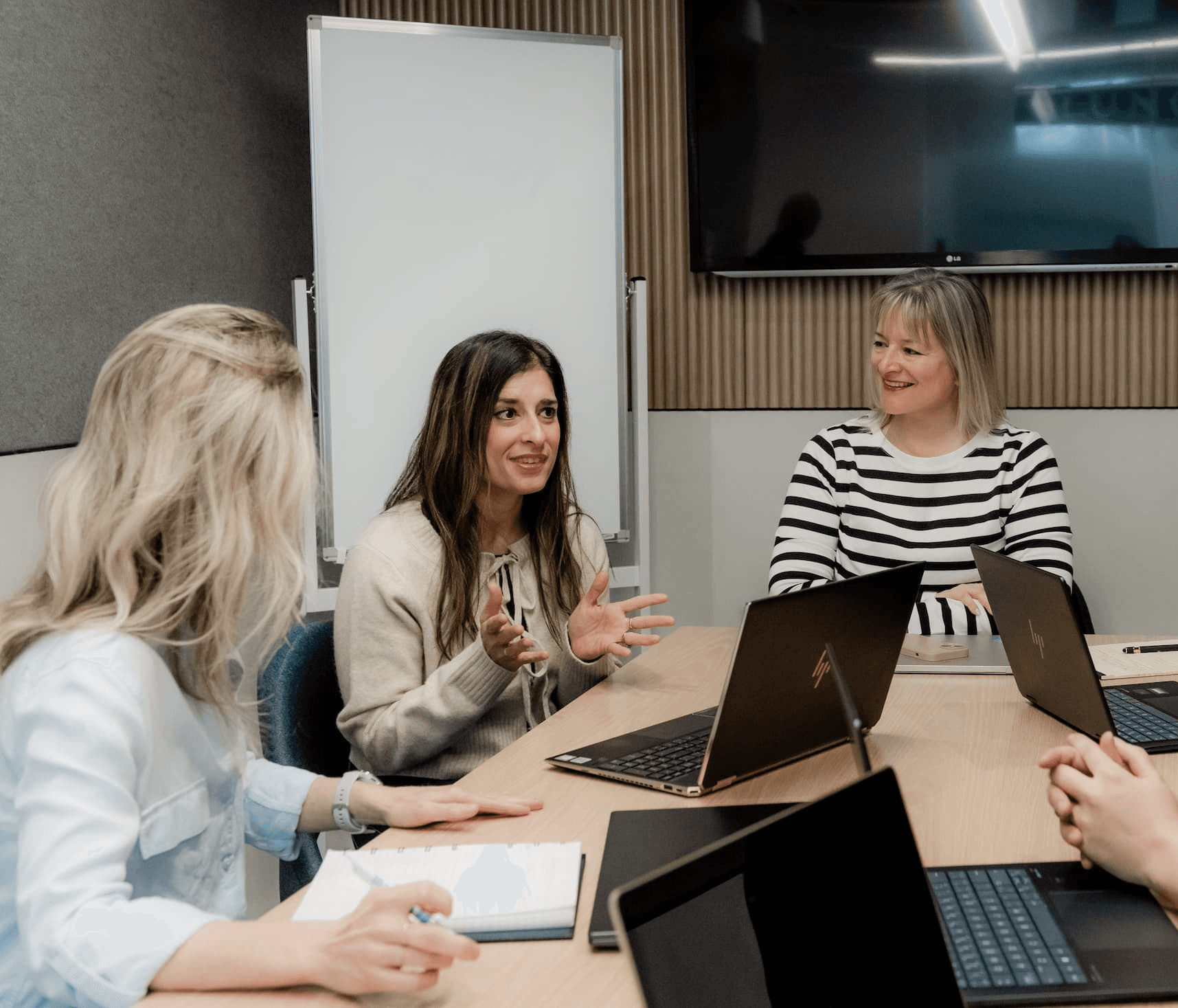 Team meeting with three women around a table, one speaking while gesturing, others listening with laptops open.