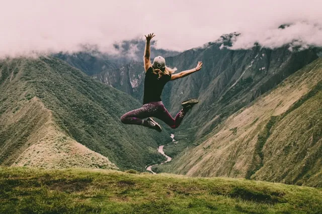 Woman jumping with arms raised on a mountain cliff overlooking a scenic valley.