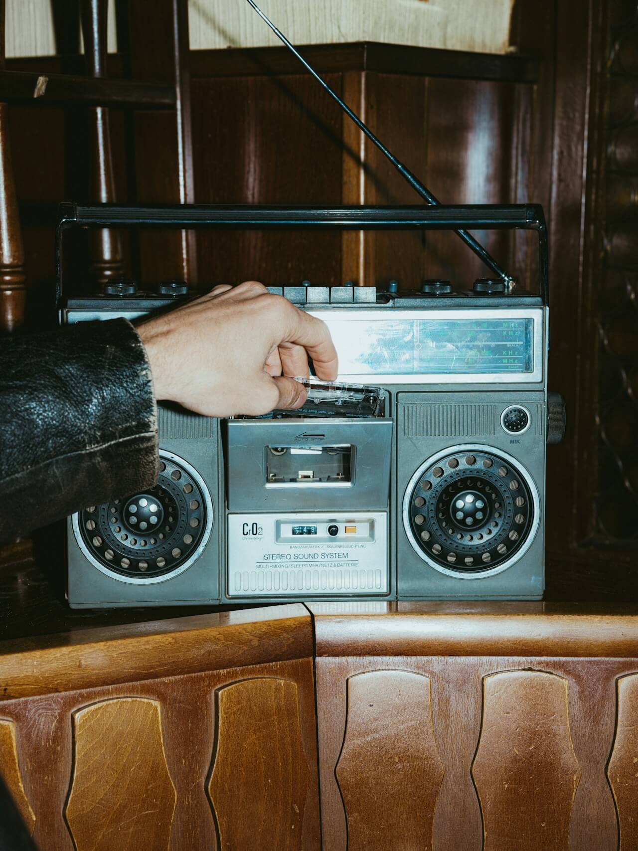 A hand in a leather jacket is inserting a cassette into a vintage boombox on a wooden shelf, evoking nostalgia and retro vibes.