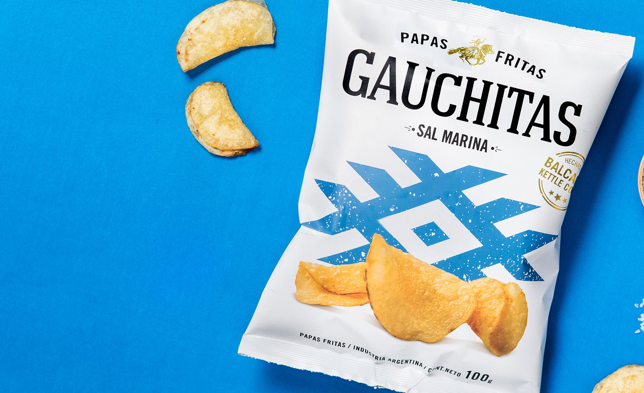 A bag of Gauchitas sea salt potato chips lies on a blue background. Three chips are scattered nearby, and the packaging features a blue geometric design.