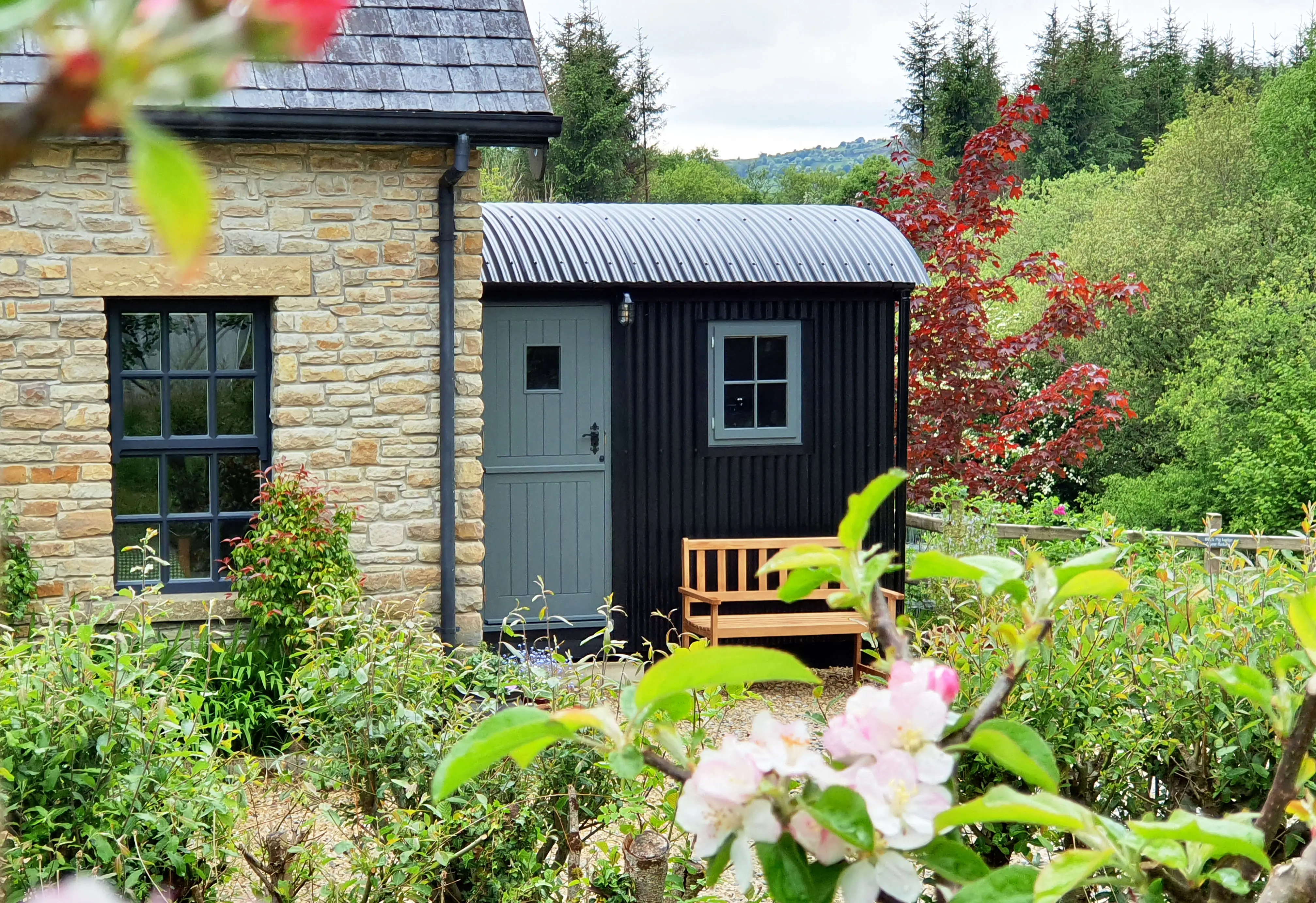 A small, modern black cabin next to a stone house, surrounded by greenery and colorful flowers.