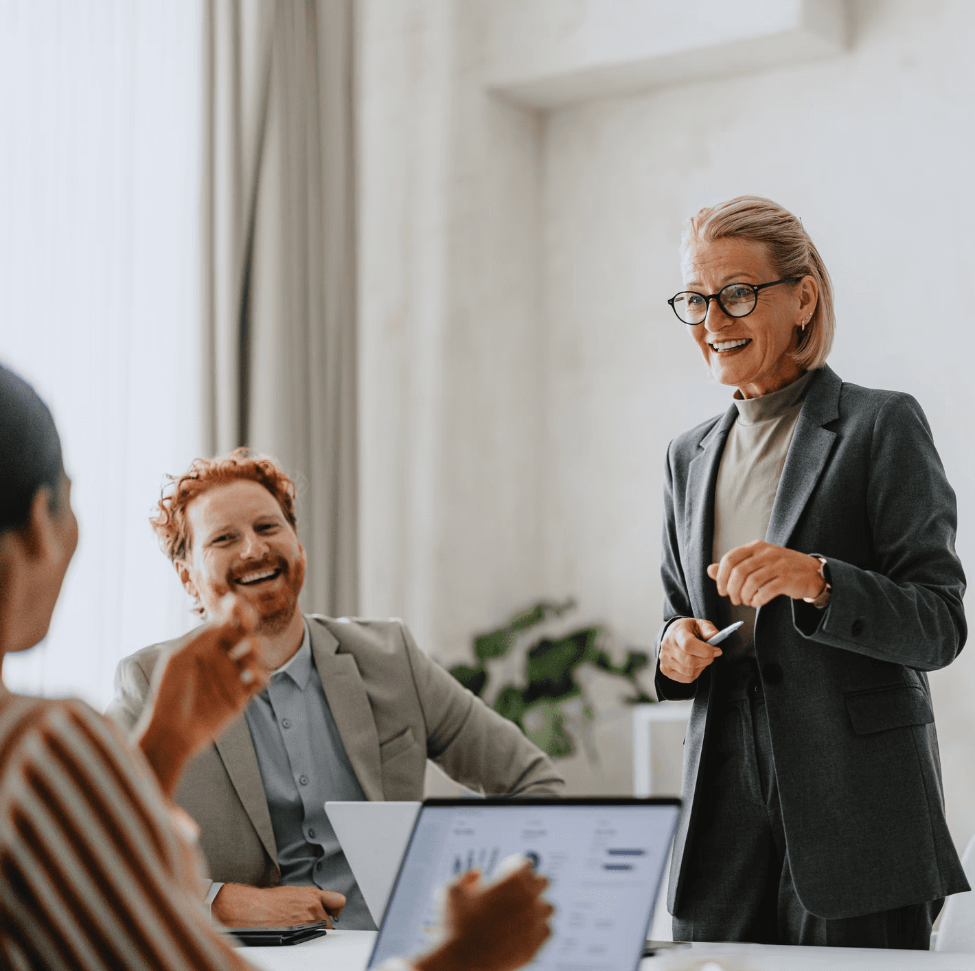 A man wearing glasses and a blazer working at a desk, writing in a notebook beside a computer monitor in a modern office.