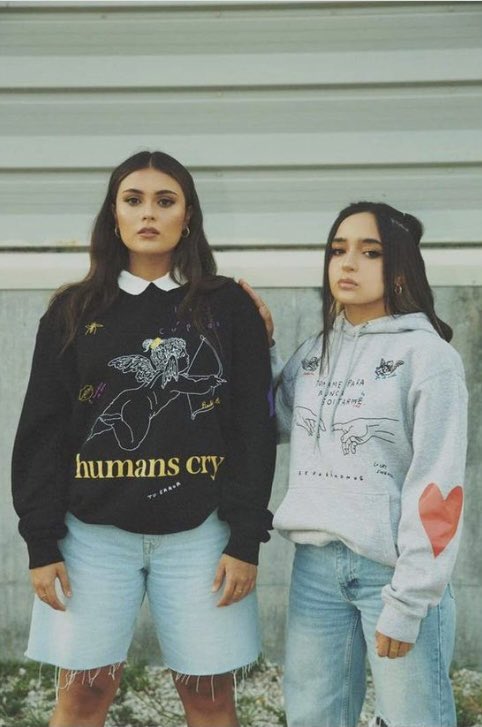 Two individuals stand together, sporting casual outfits against a simple background.