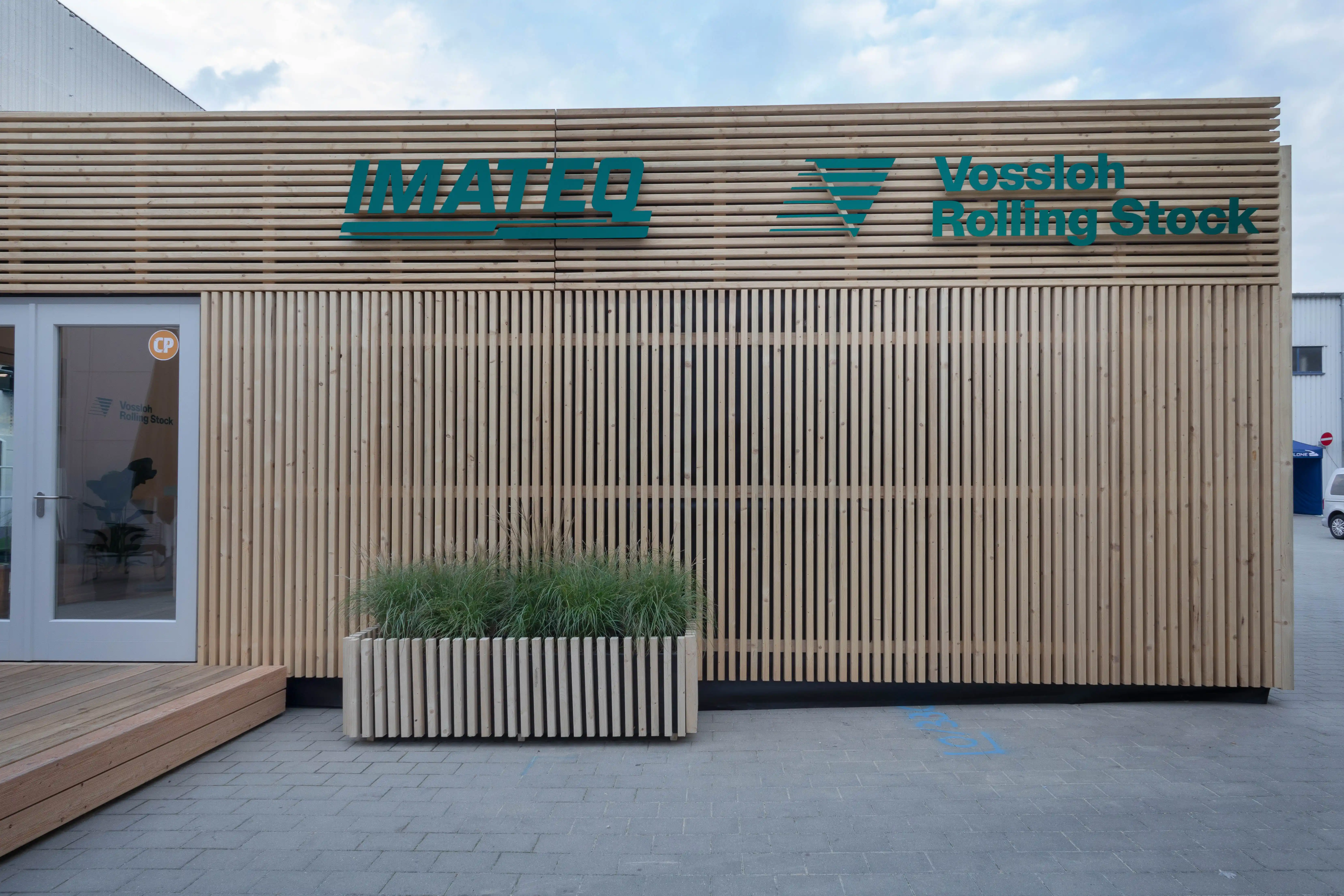 Wood-slat facade of the trade fair pavilion with green IMATEQ and Vossloh Rolling Stock logos and grass planter.