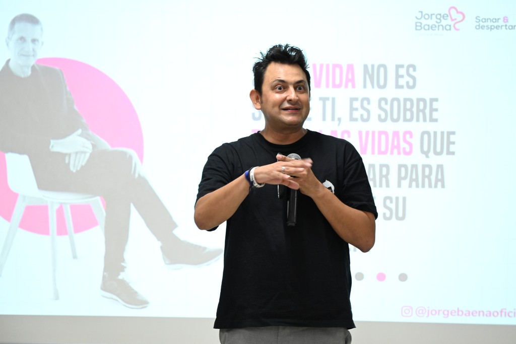 Jorge Baena de pie frente a una pantalla con una presentación colorida, sosteniendo un micrófono y mirando al público durante una conferencia.