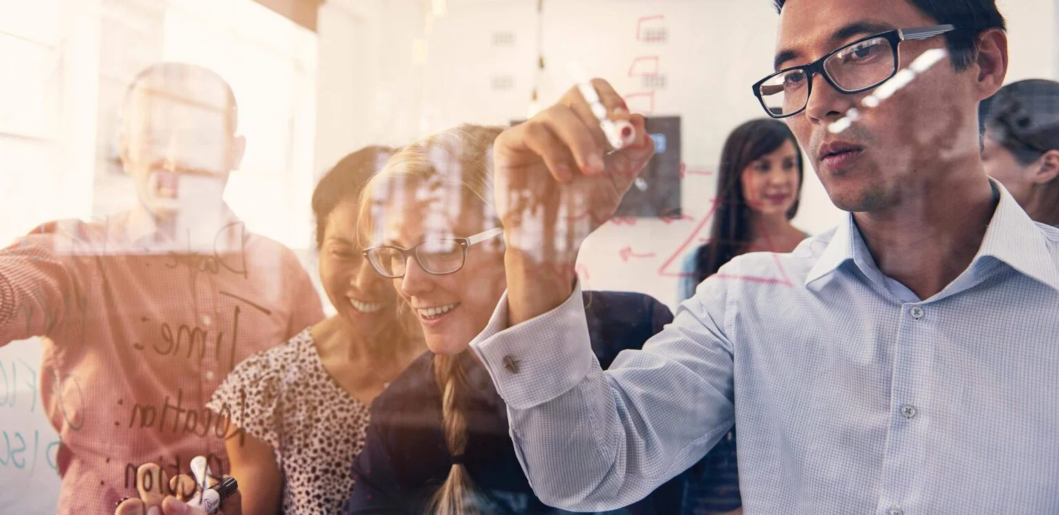 Colleagues smiling and sketching ideas on a glass wall, a man in glasses drawing in the foreground