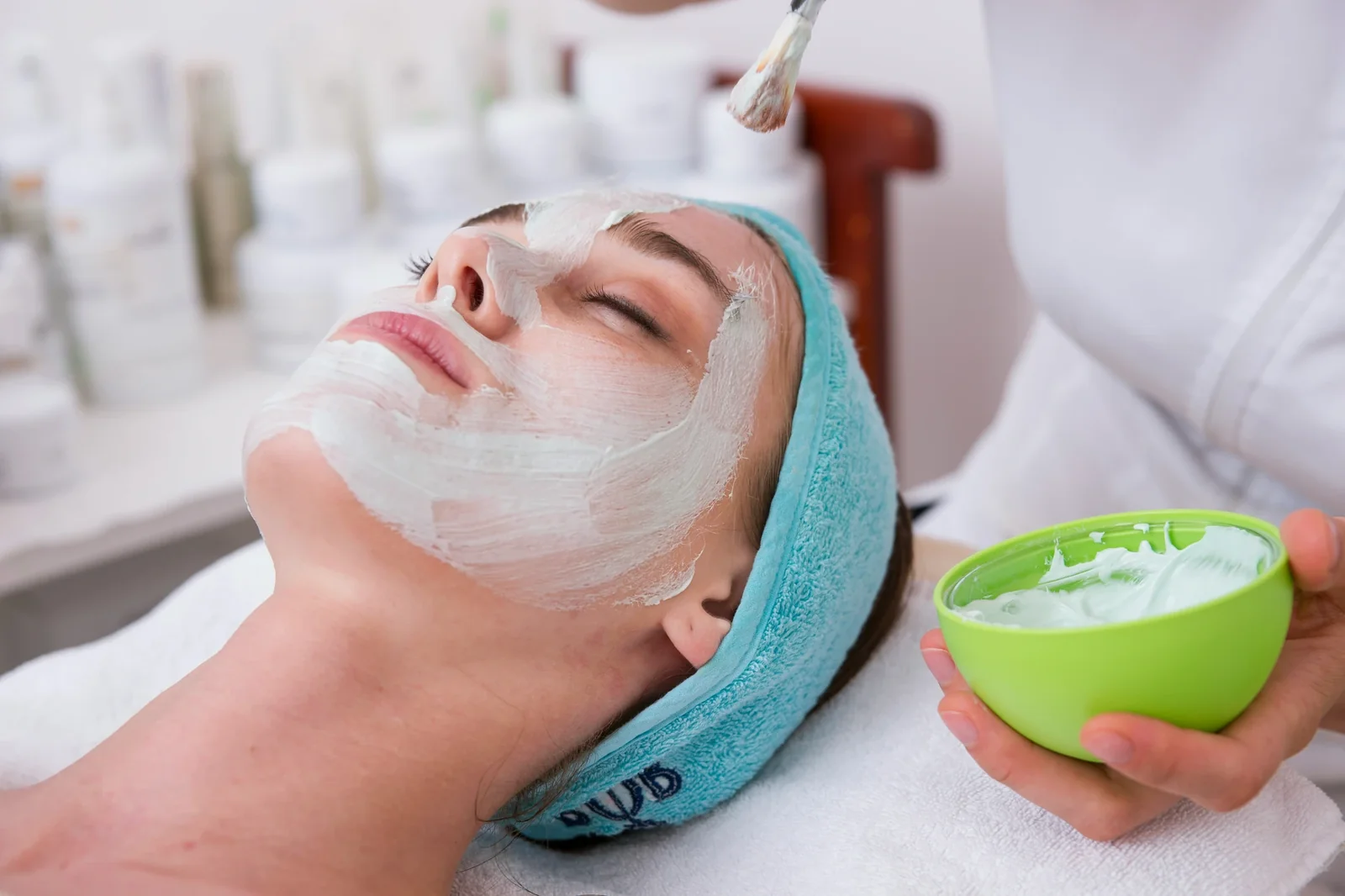 Esthetician applies white facial mask to relaxed client in spa setting with green bowl of product.