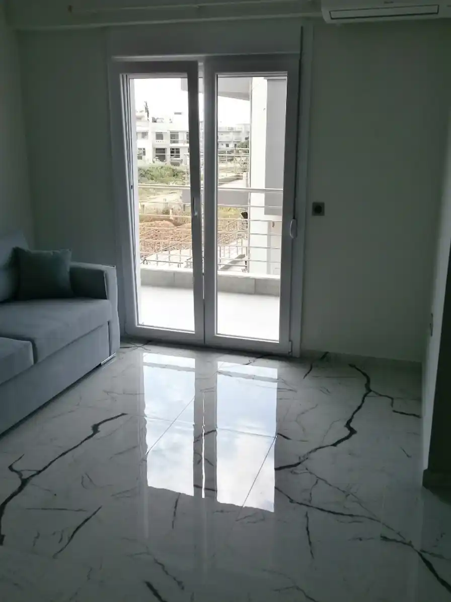 Μοντέρνο σαλόνι με γκρι καναπέ, μαρμάρινο δάπεδο και μεγάλη μπαλκονόπορτα που οδηγεί σε βεράντα.
