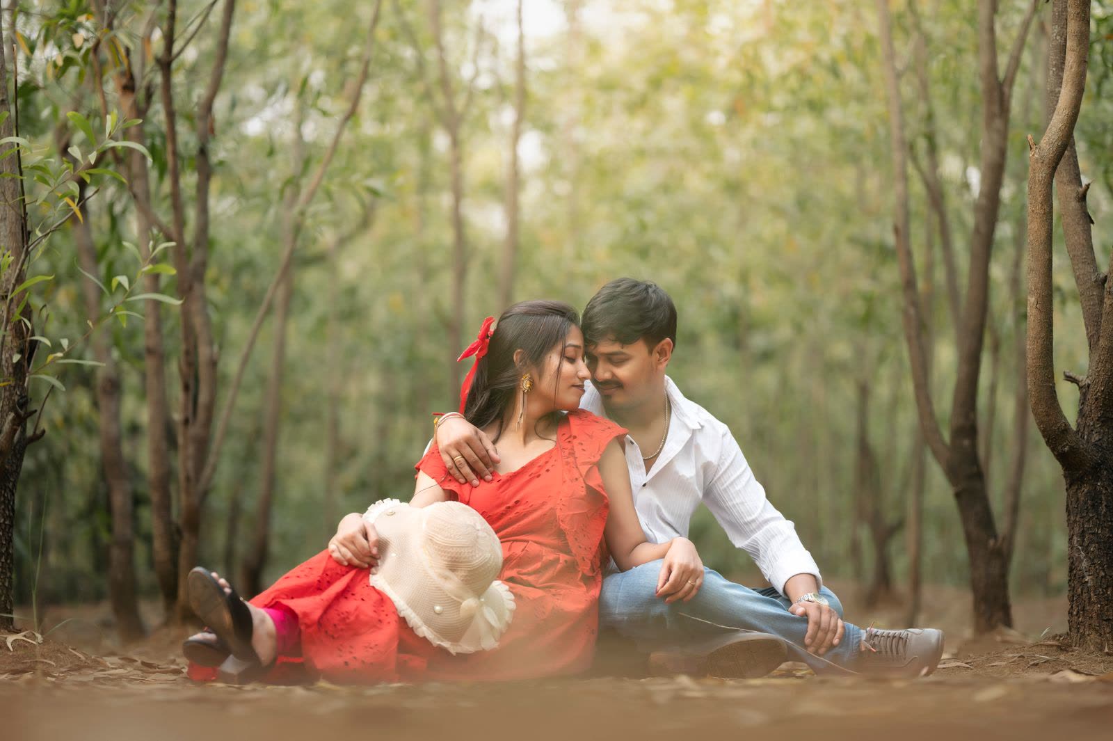 Couple sitting on the ground in a forest, the woman in red holding a straw hat
