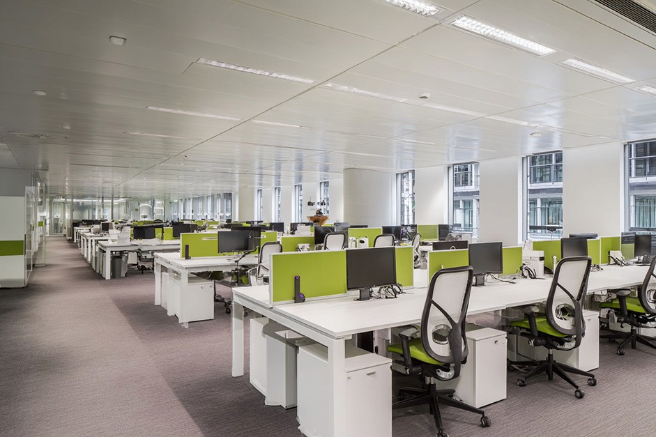 A modern, spacious open-plan office features rows of white desks equipped with dual monitors and ergonomic chairs, set against large windows that provide natural light and views of an urban environment.