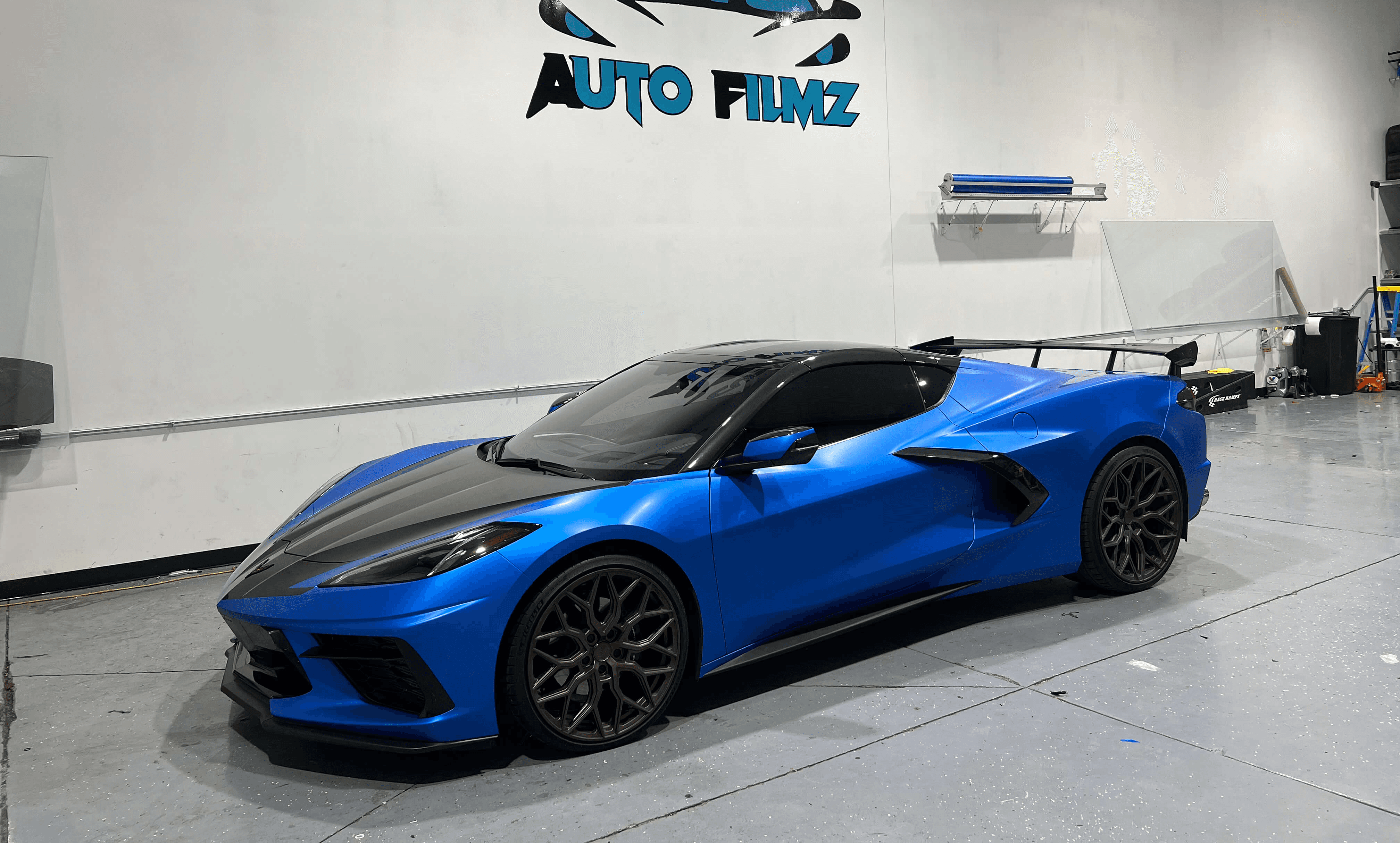 A vibrant blue Chevrolet Corvette C8 with a black hood and bronze wheels is parked indoors on a polished gray floor.