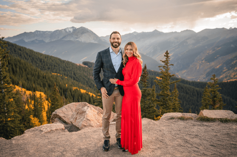 Couple in a red dress and blue jacket posing on a rocky mountain overlook at sunset