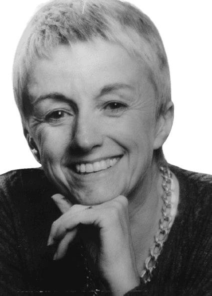 A black-and-white portrait of a smiling woman with short, cropped hair, resting her chin on her hand in a relaxed, thoughtful pose. She wears a chunky chain necklace and a dark top, with a friendly, approachable expression that highlights her cheerful demeanor. The simple background keeps the focus on her expressive face and warm smile.