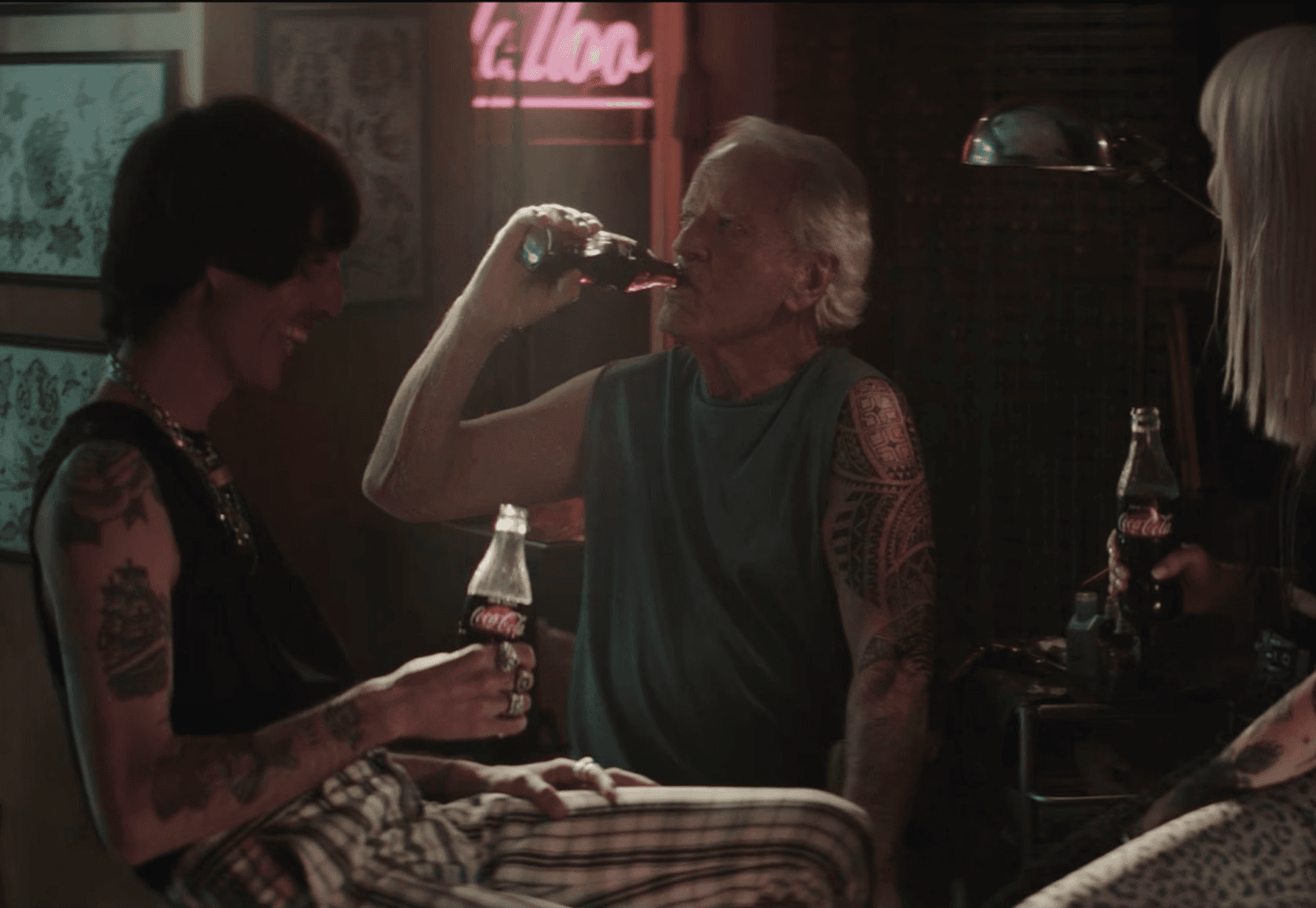 Three tattooed people sharing Coca-Cola glass bottles in moody neon-lit tattoo parlour, older man with sleeve tattoo drinking Coke flanked by two women, cinematic TV advertisement still