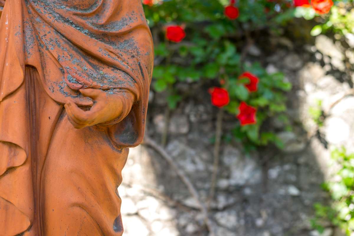 A terracotta-colored statue draped in robes standing before a stone wall with red flowers and green leaves.