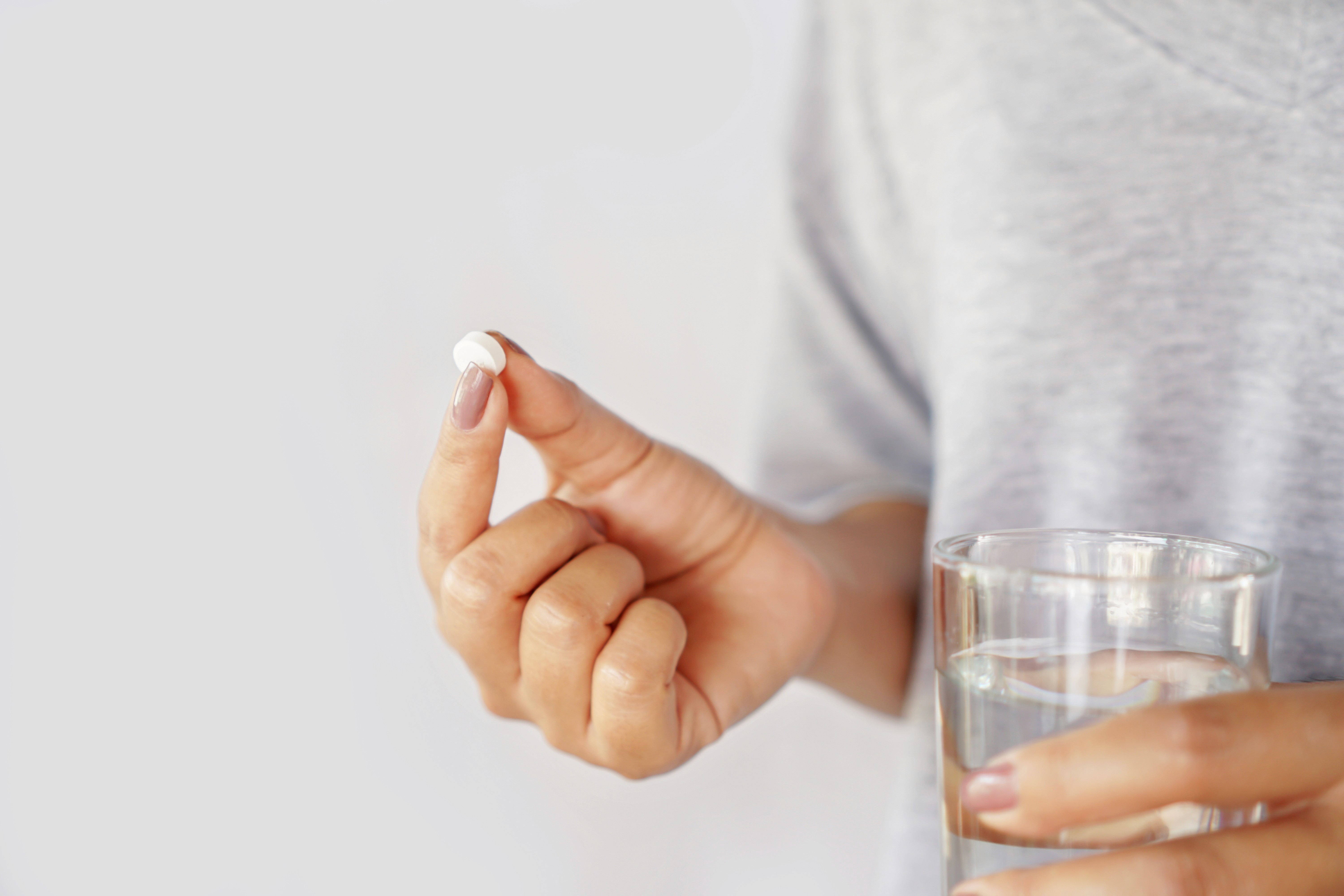 Black and white image of a person holding a small white pill between their fingers next to a glass of water.