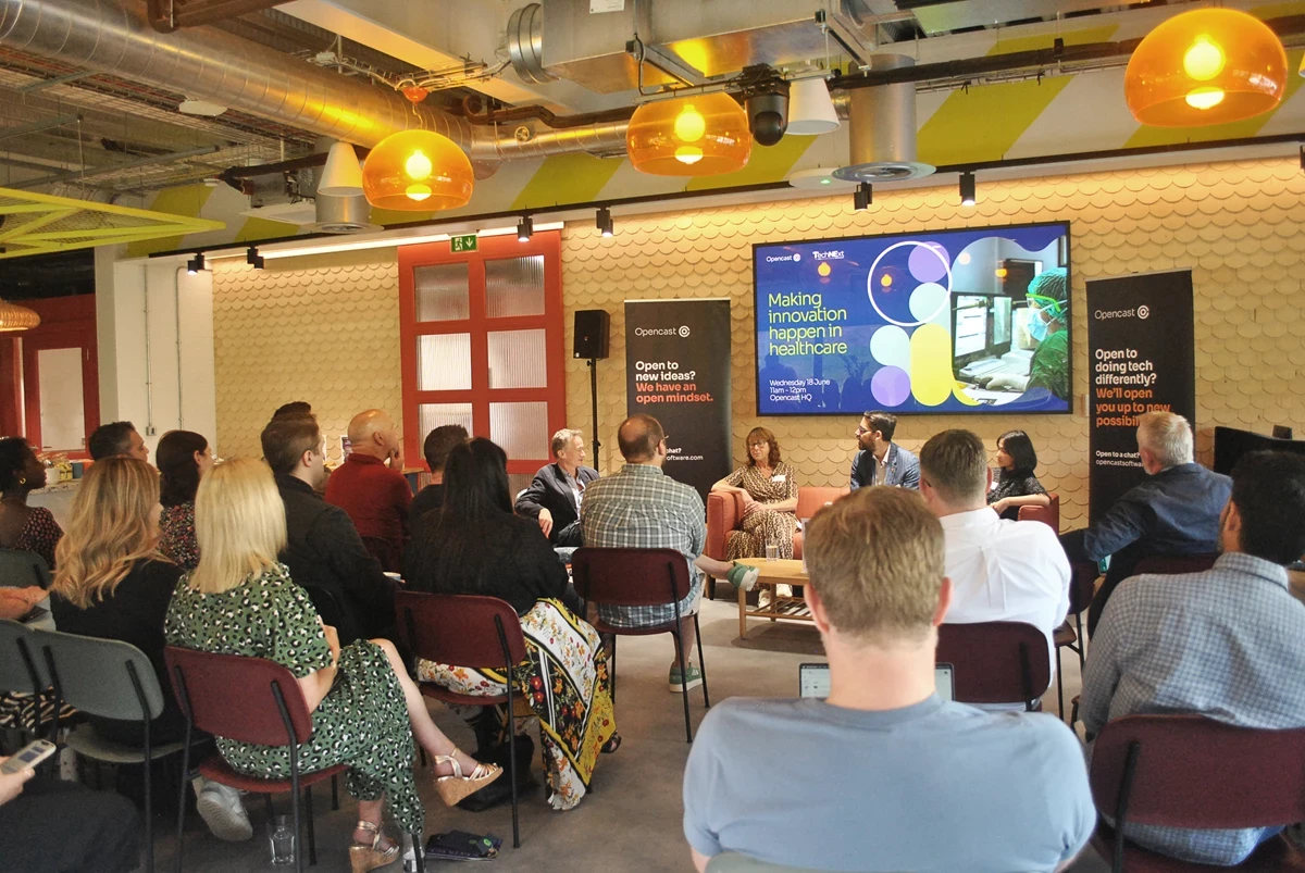 Audience watching a panel discussion in a modern room with Opencast banners and a screen about healthcare innovation.