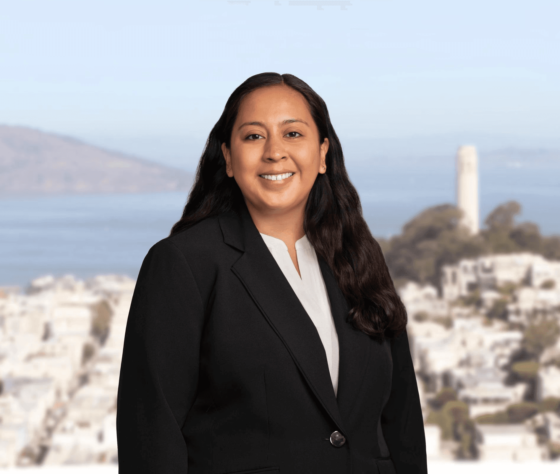 Uma mulher vestindo um blazer preto e uma blusa branca está sorrindo diante de um cenário urbano desfocado, com uma torre alta em destaque e uma vista ao longe da água e de uma paisagem montanhosa, transmitindo uma postura profissional e acessível.