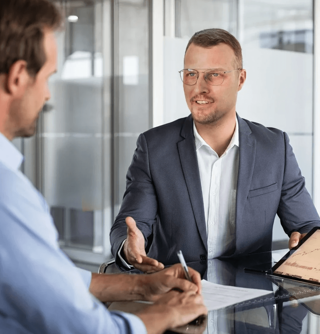 Zwei Geschäftspartner in einem Besprechungsraum; ein Mann mit Brille im Anzug führt ein Gespräch, während sein Gegenüber sich Notizen macht und ein Tablet auf dem Tisch liegt.