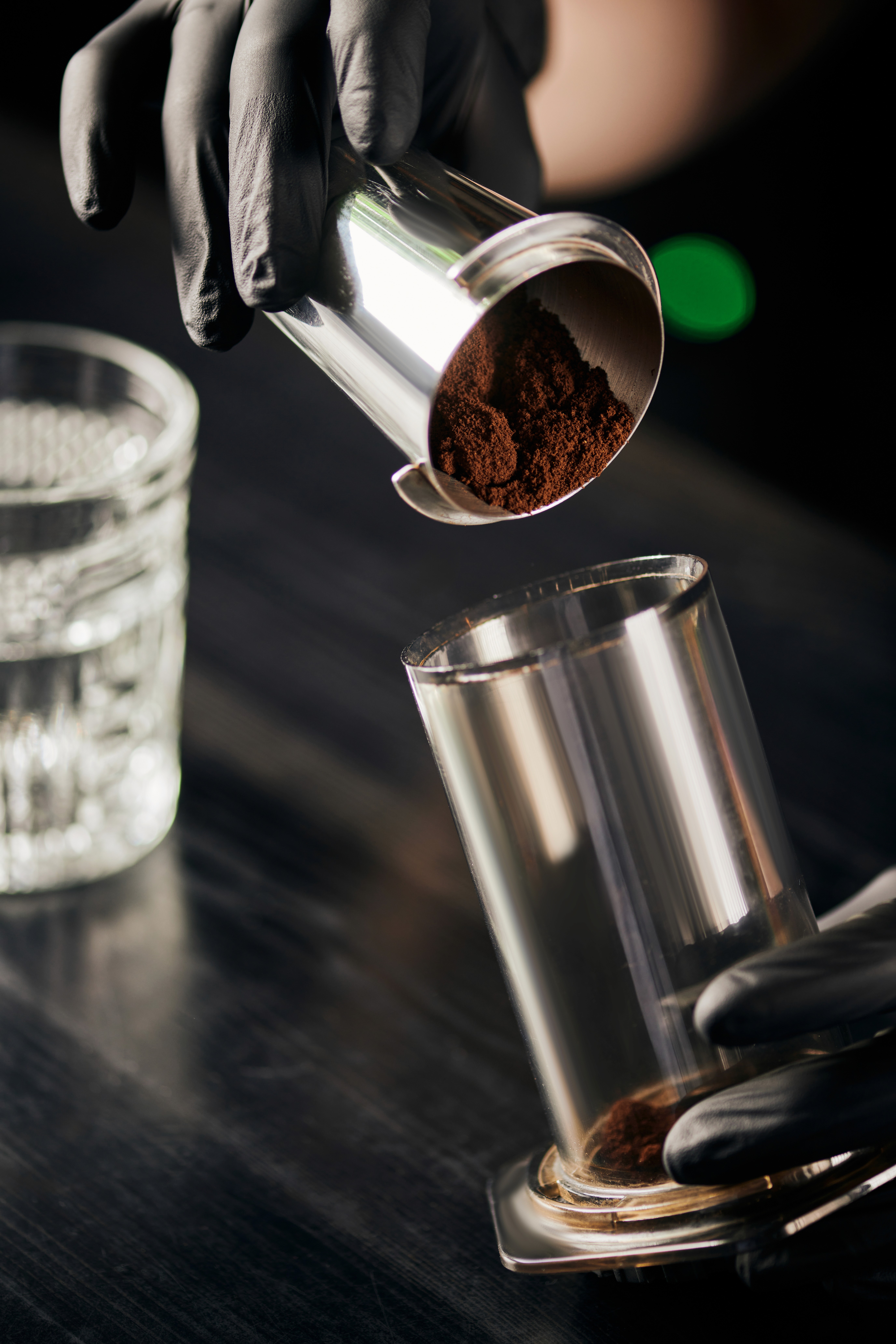 Barista pouring freshly ground coffee from a metal dosing cup into a grinder container, captured in a dark, moody café close-up.