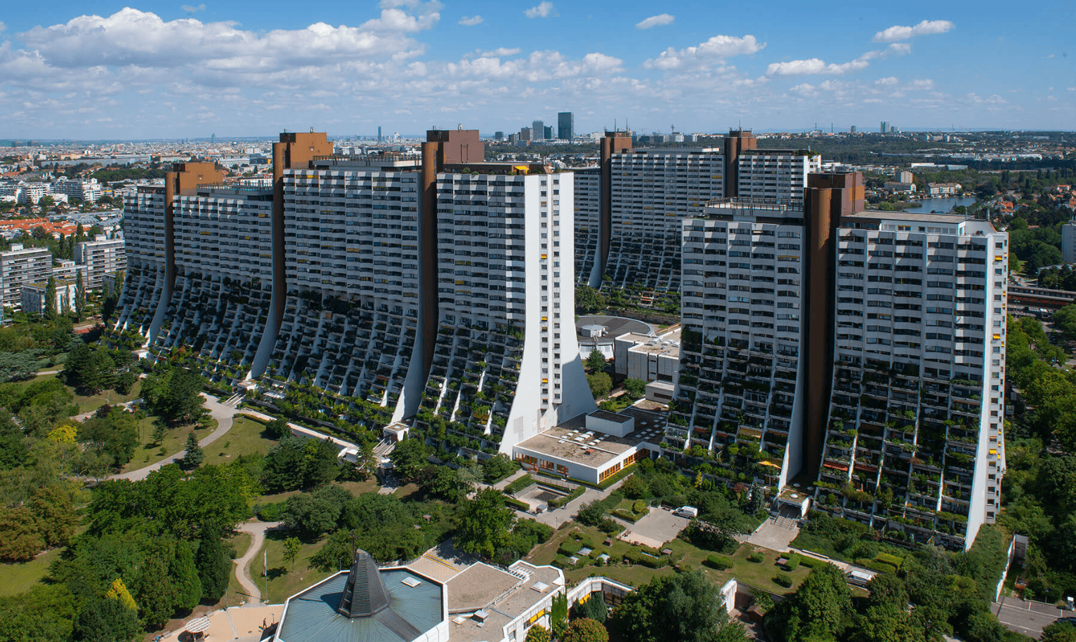 Aerial view of the curved, terraced high-rise residential buildings of the Alt Erlaa Wohnpark situated among green parkland with a city skyline in the background under a partly cloudy blue sky.