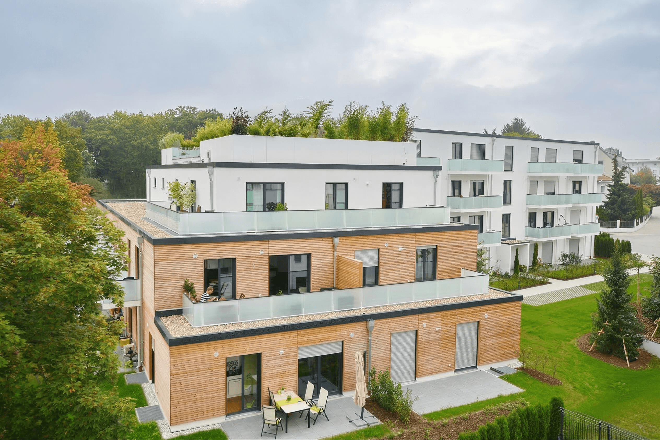 Moderne Wohnanlage mit Holzelementen, Terrassen und viel Grün, im Hintergrund Baumallee und weitere Gebäude.